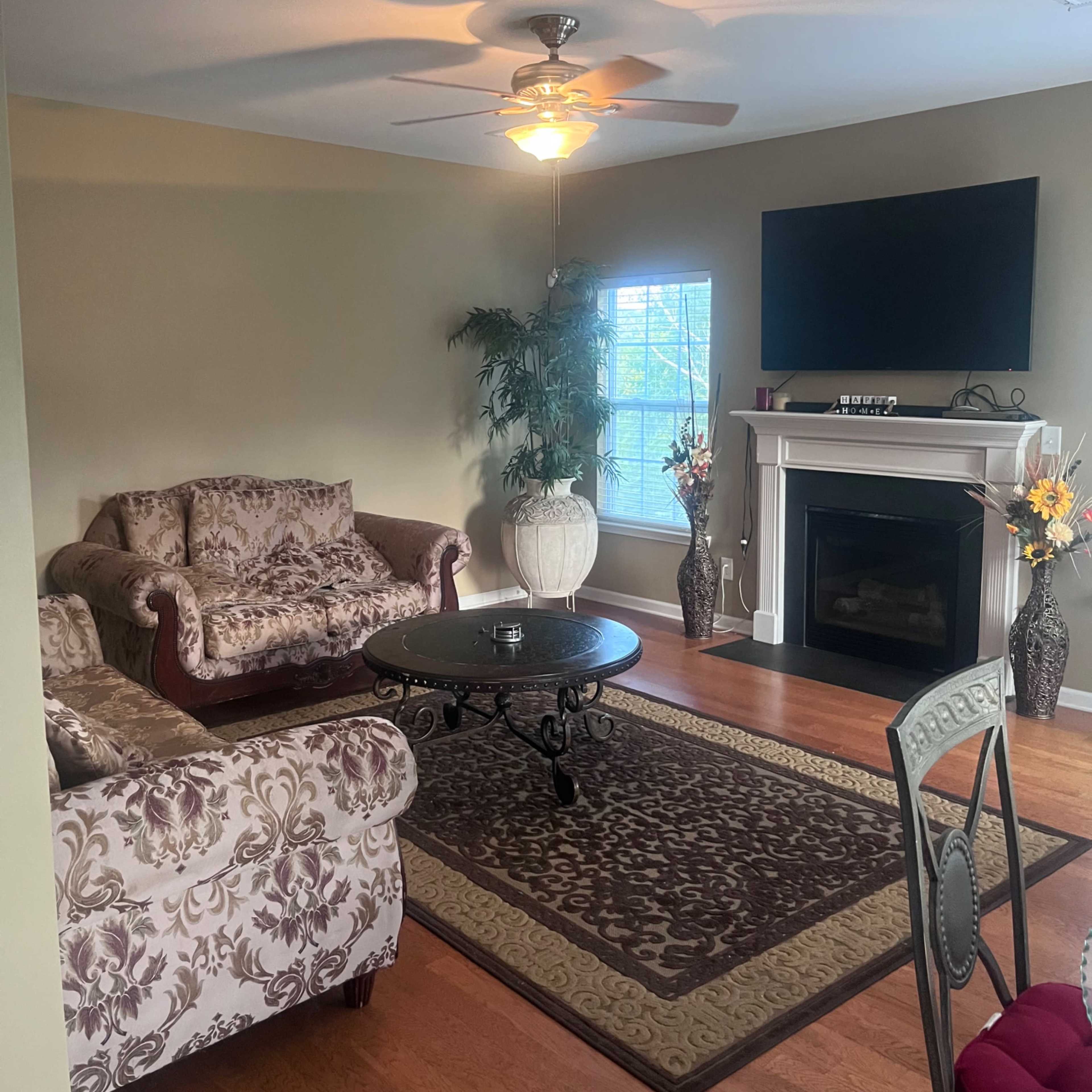 The living room features a floral-patterned couch and loveseat, a round coffee table, a fireplace with a television above it, and large windows allowing natural light to enter.