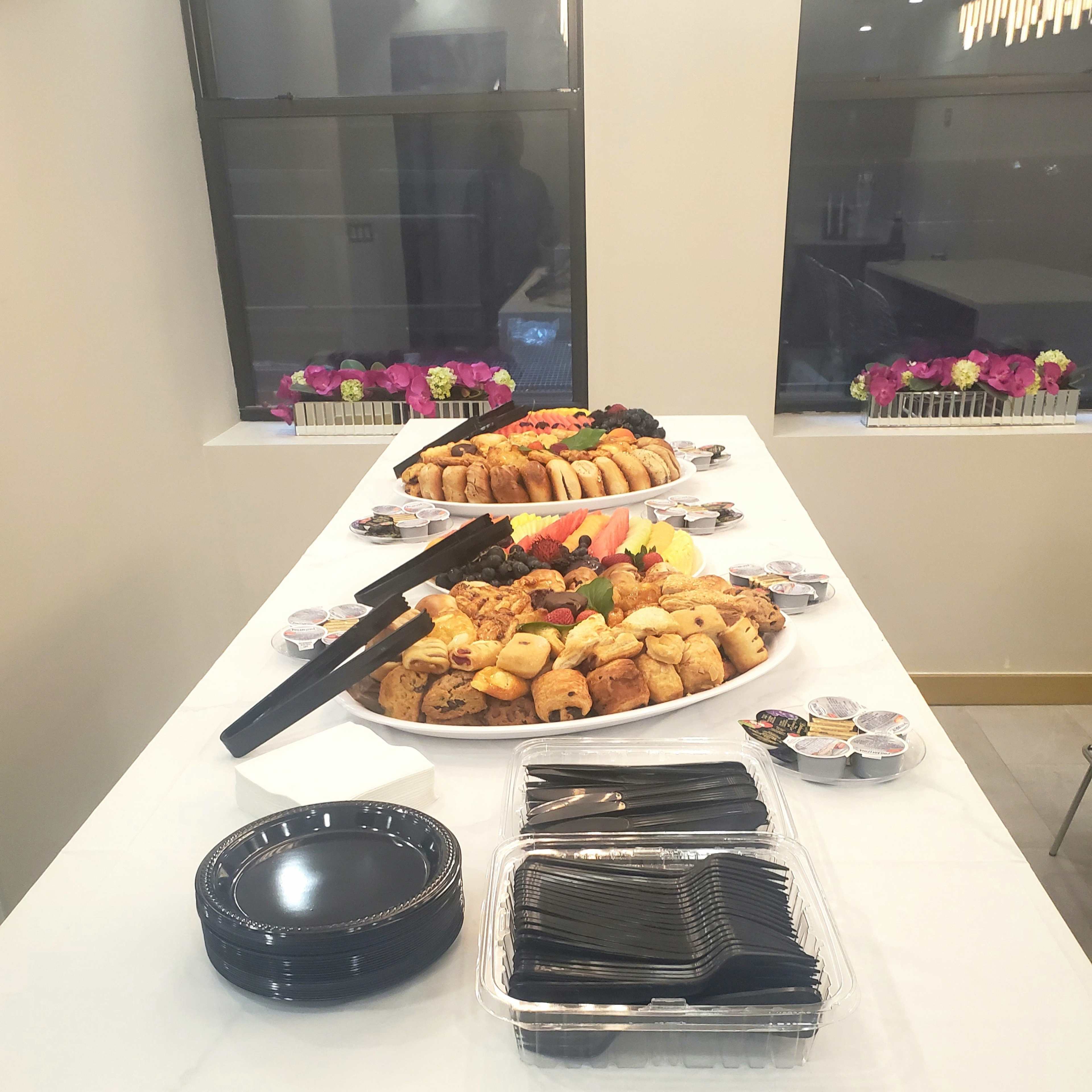 A table is set with a variety of food platters, including fruit, pastries, and small dishes, alongside black plates and utensils.