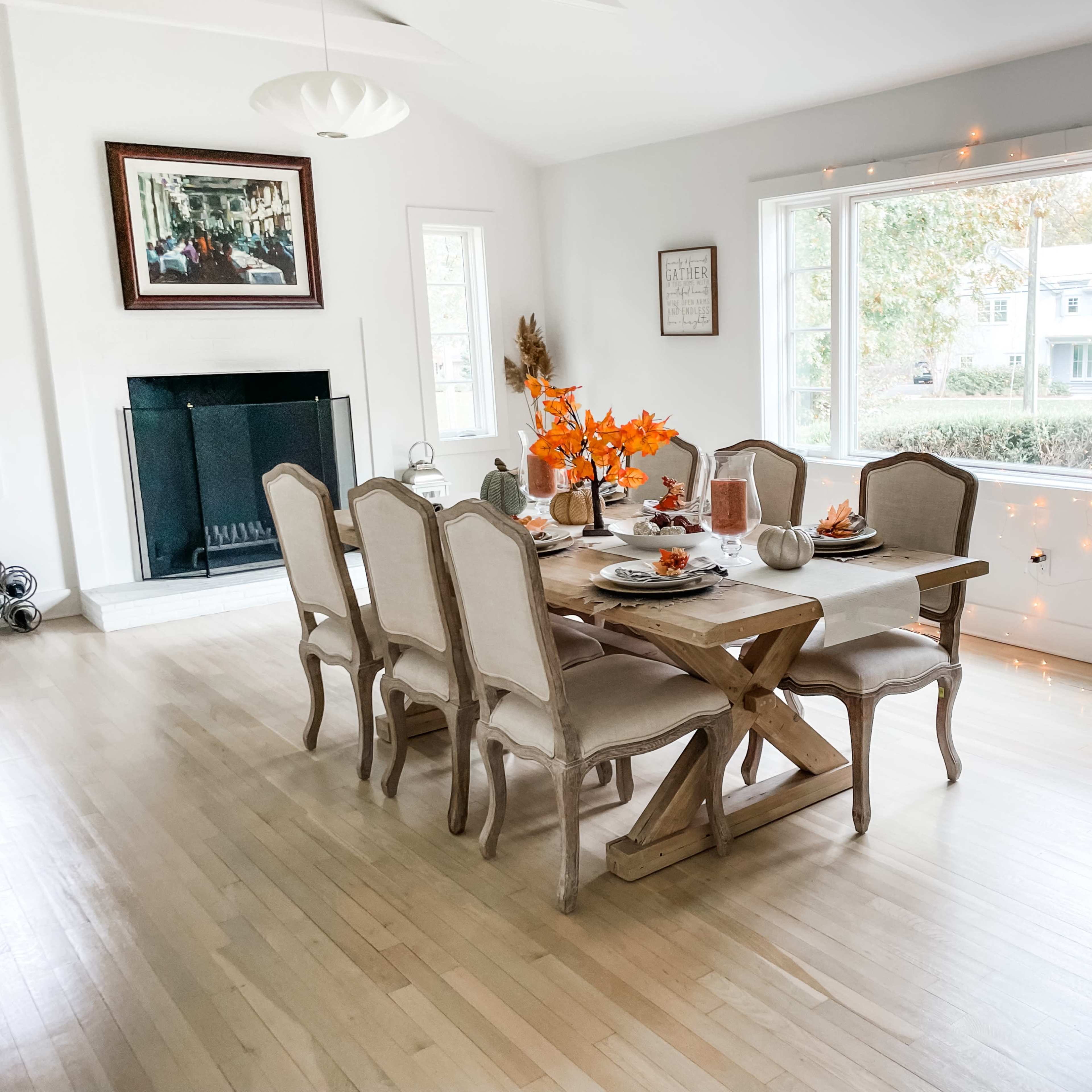 A dining room features a large wooden table set for a meal, surrounded by chairs, with orange autumn decorations and a view of a tree-lined street through the window.