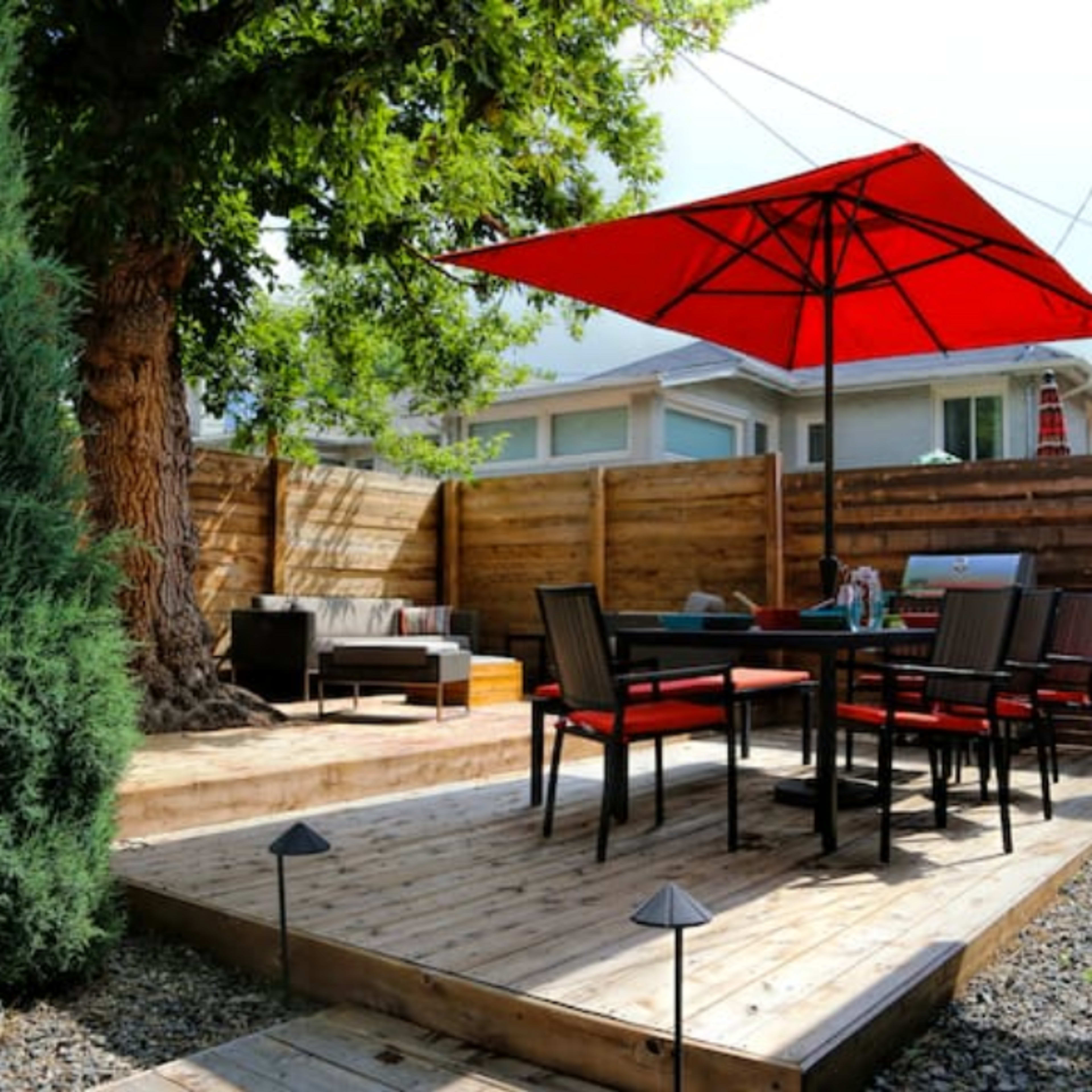 A wooden patio features a table and chairs under a red umbrella, surrounded by wooden fencing and greenery.