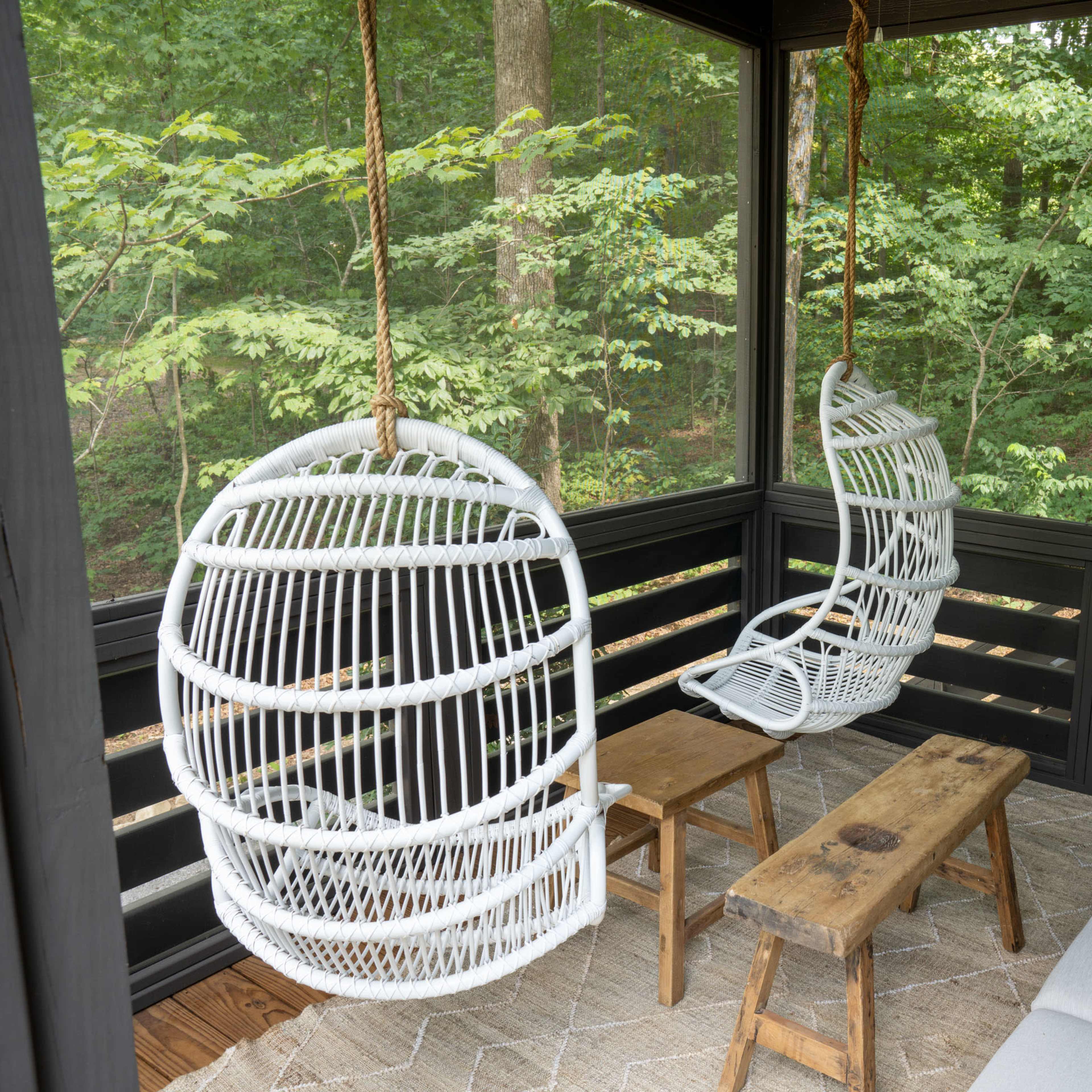A screened porch with two hanging white wicker chairs, a wooden coffee table, and a sectional sofa, surrounded by greenery.