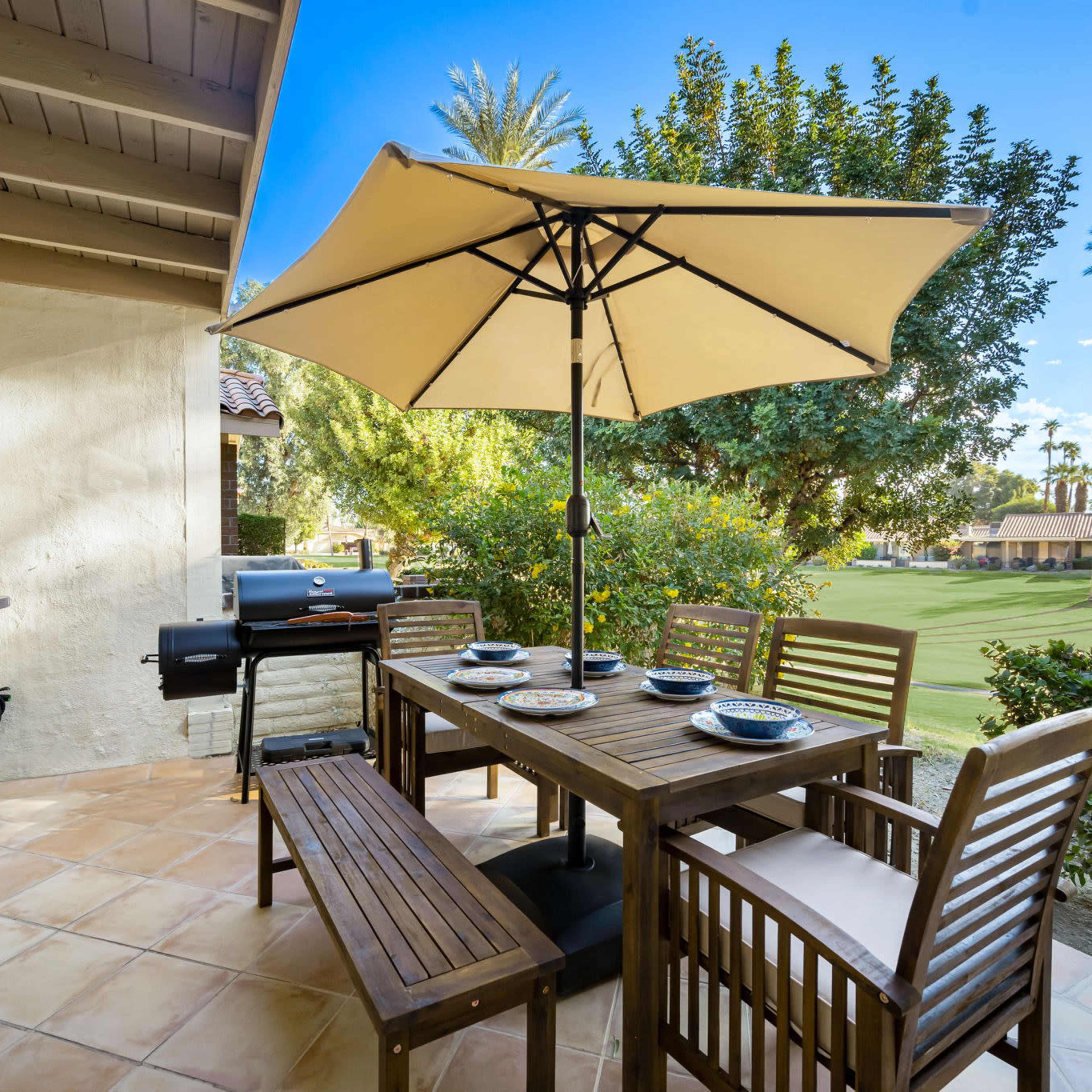 A patio with a wooden dining table and chairs, an umbrella for shade, and a grill, surrounded by a view of palm trees and a green lawn.