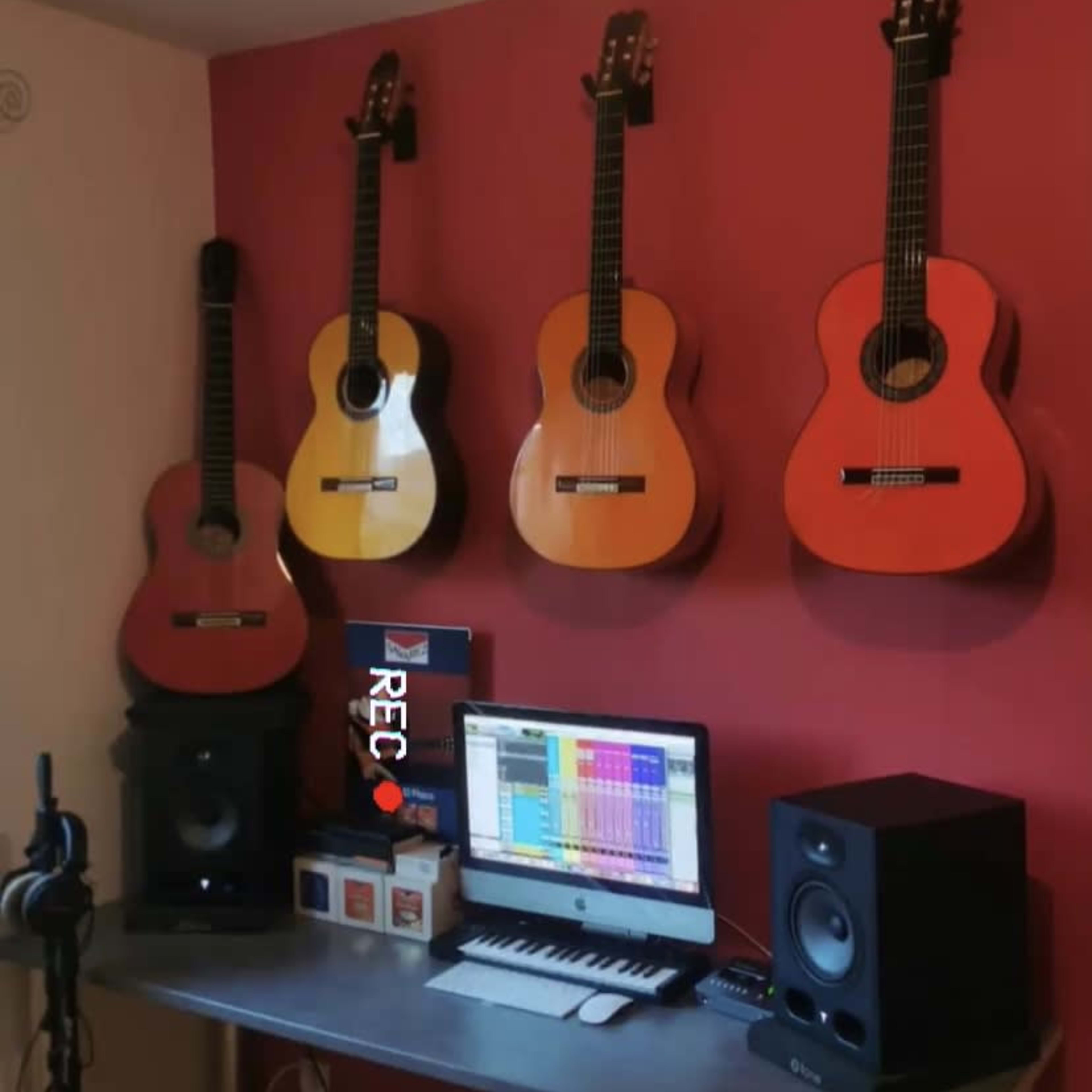 A home recording studio setup featuring four guitars mounted on a red wall, with a computer and audio equipment on a desk below.