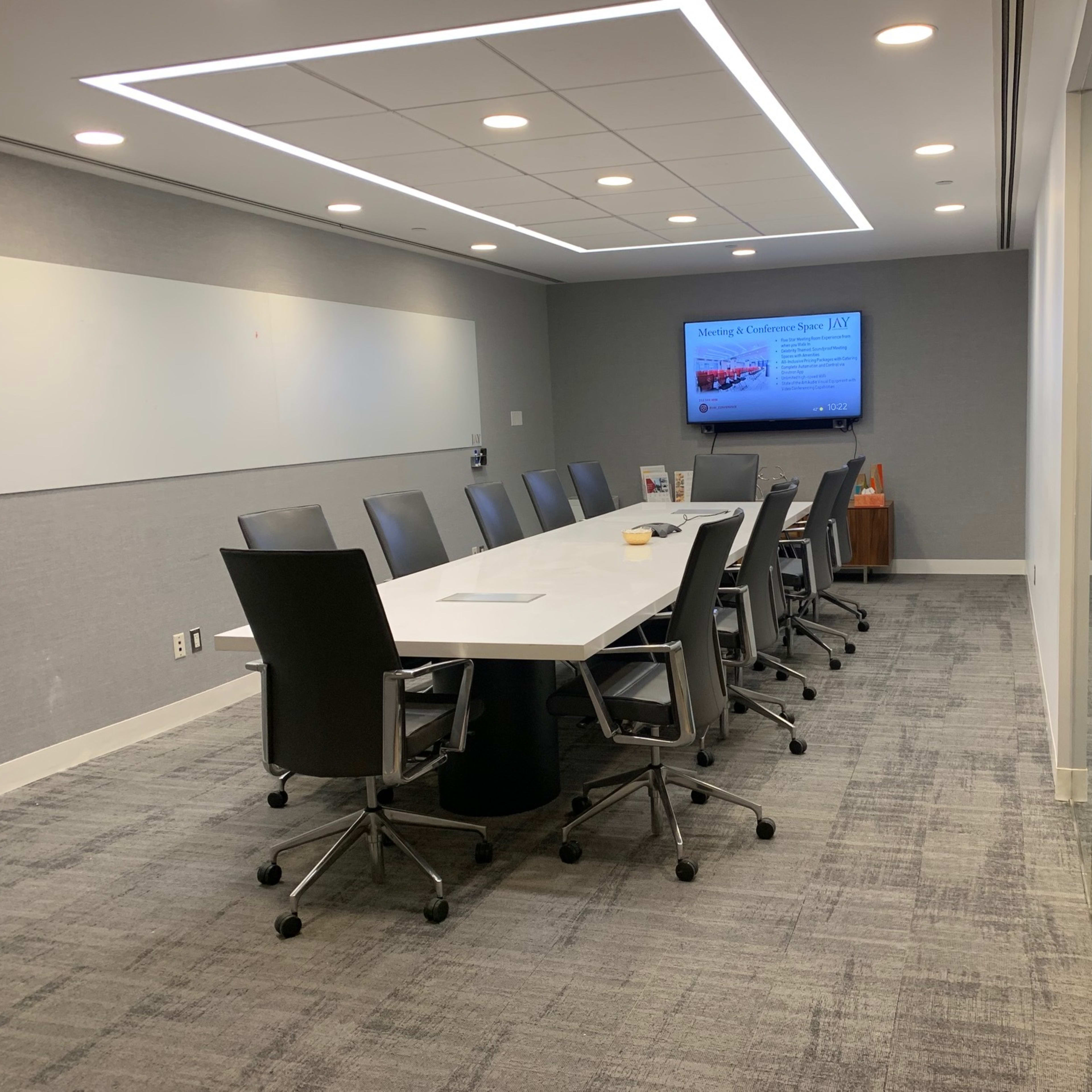 A modern conference room features a long table surrounded by black office chairs, with a screen displaying a presentation on the wall.