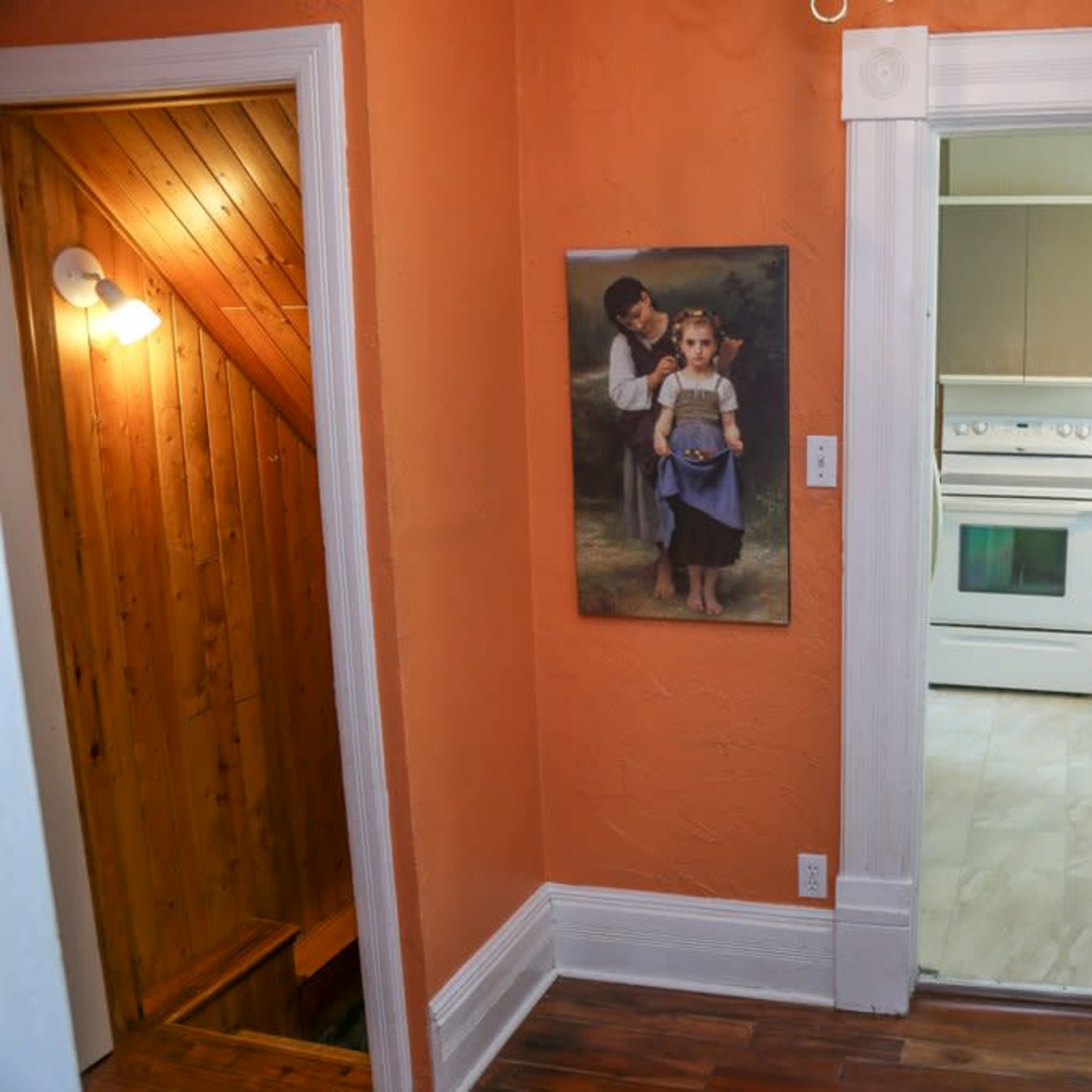 A hallway with orange walls, a staircase visible on the left, and an open doorway leading to a kitchen.