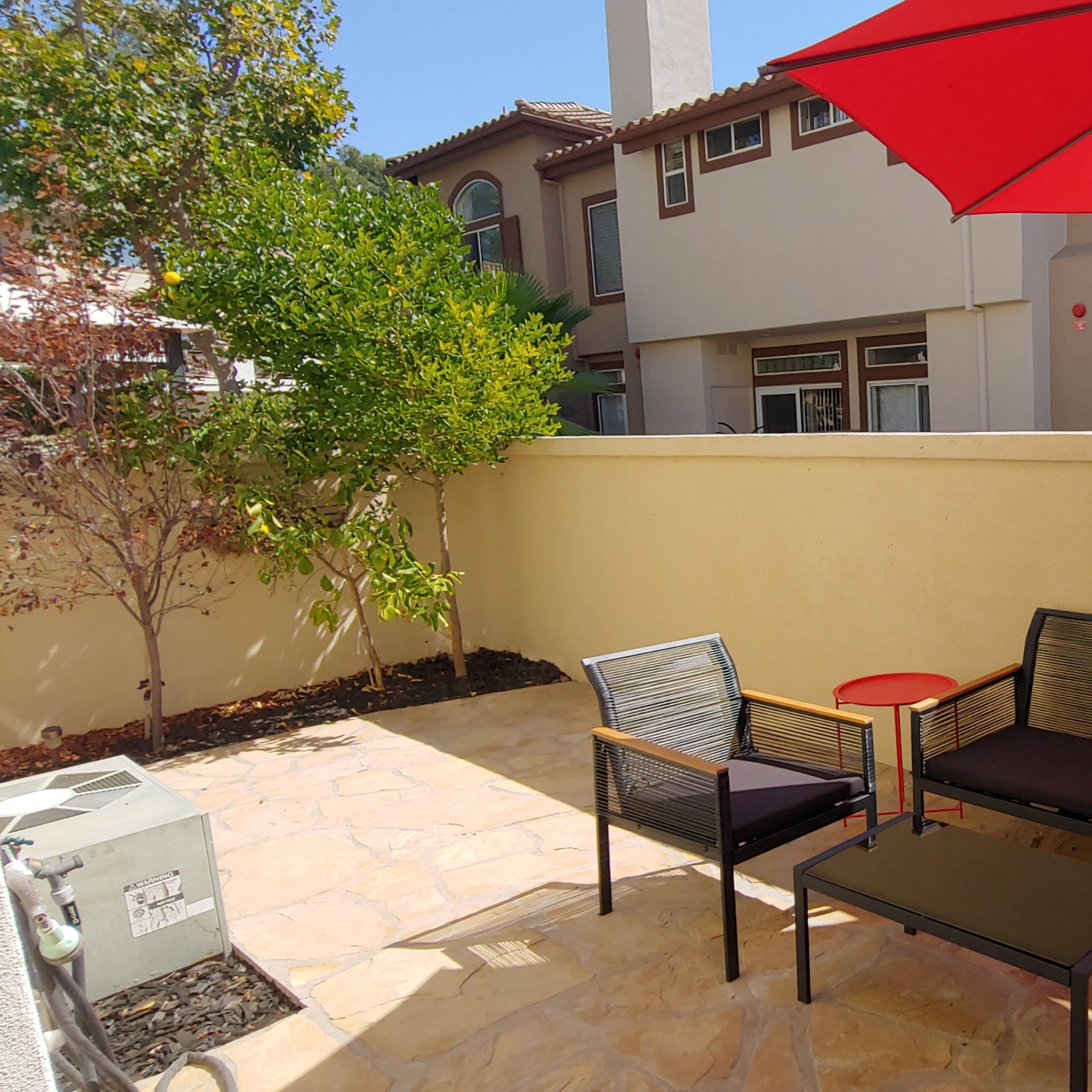 A small courtyard features two seating areas with black cushions, a red umbrella, and a stone patio surrounded by low walls and greenery.