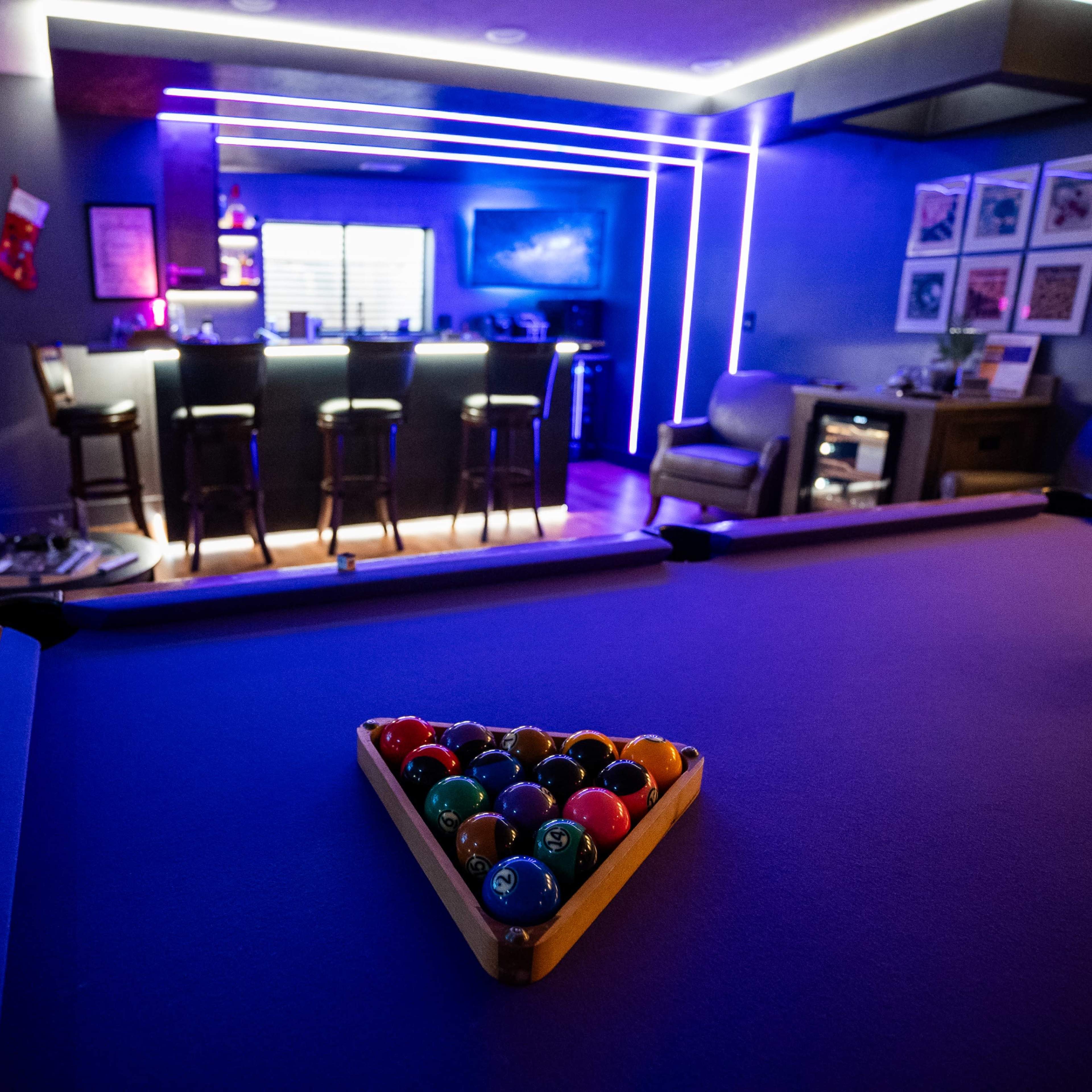 A billiard table with a triangle of colored balls is in the foreground, while a modern bar area with purple lighting is visible in the background.