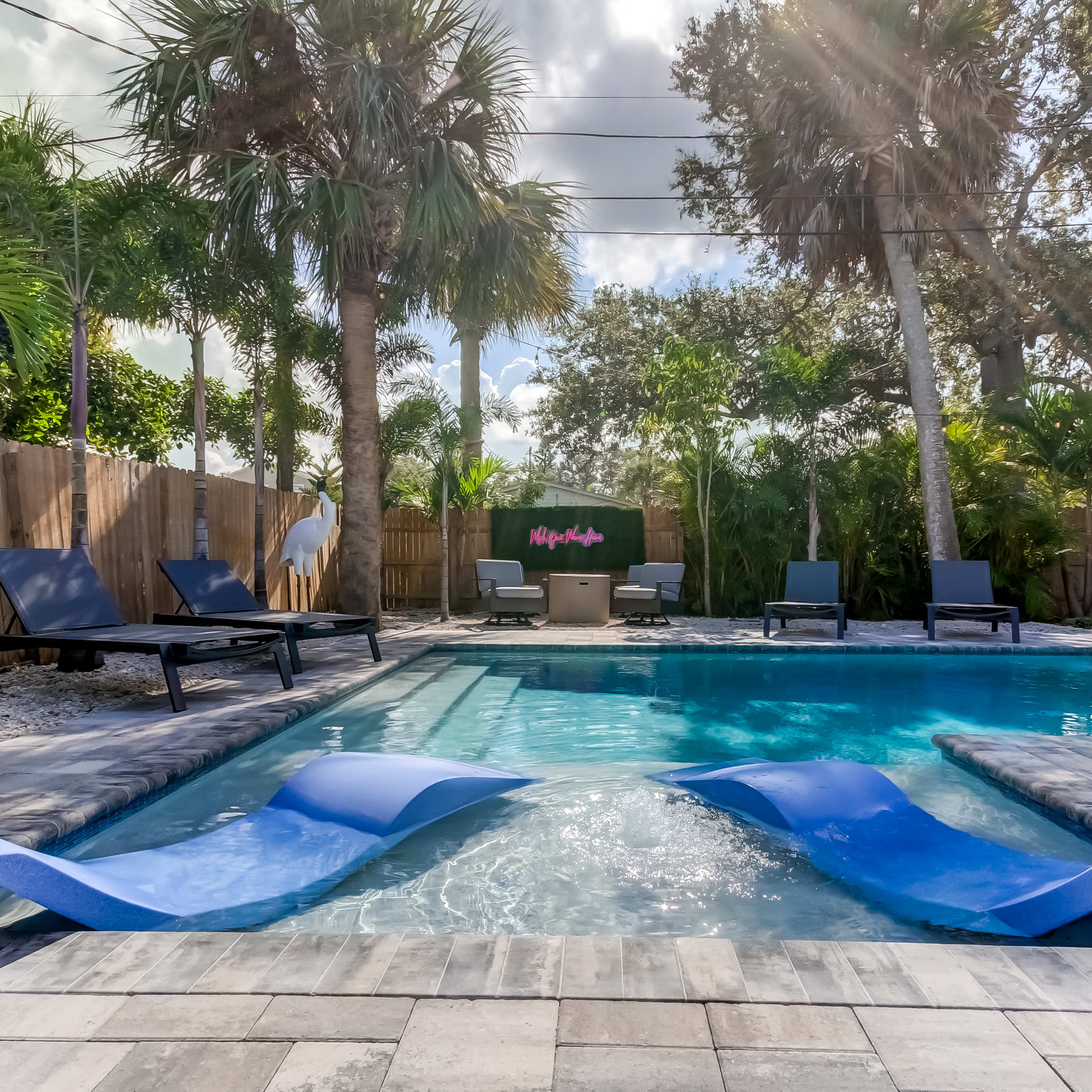 A blue swimming pool with lounge chairs and palm trees is surrounded by a stone patio and wooden fencing.