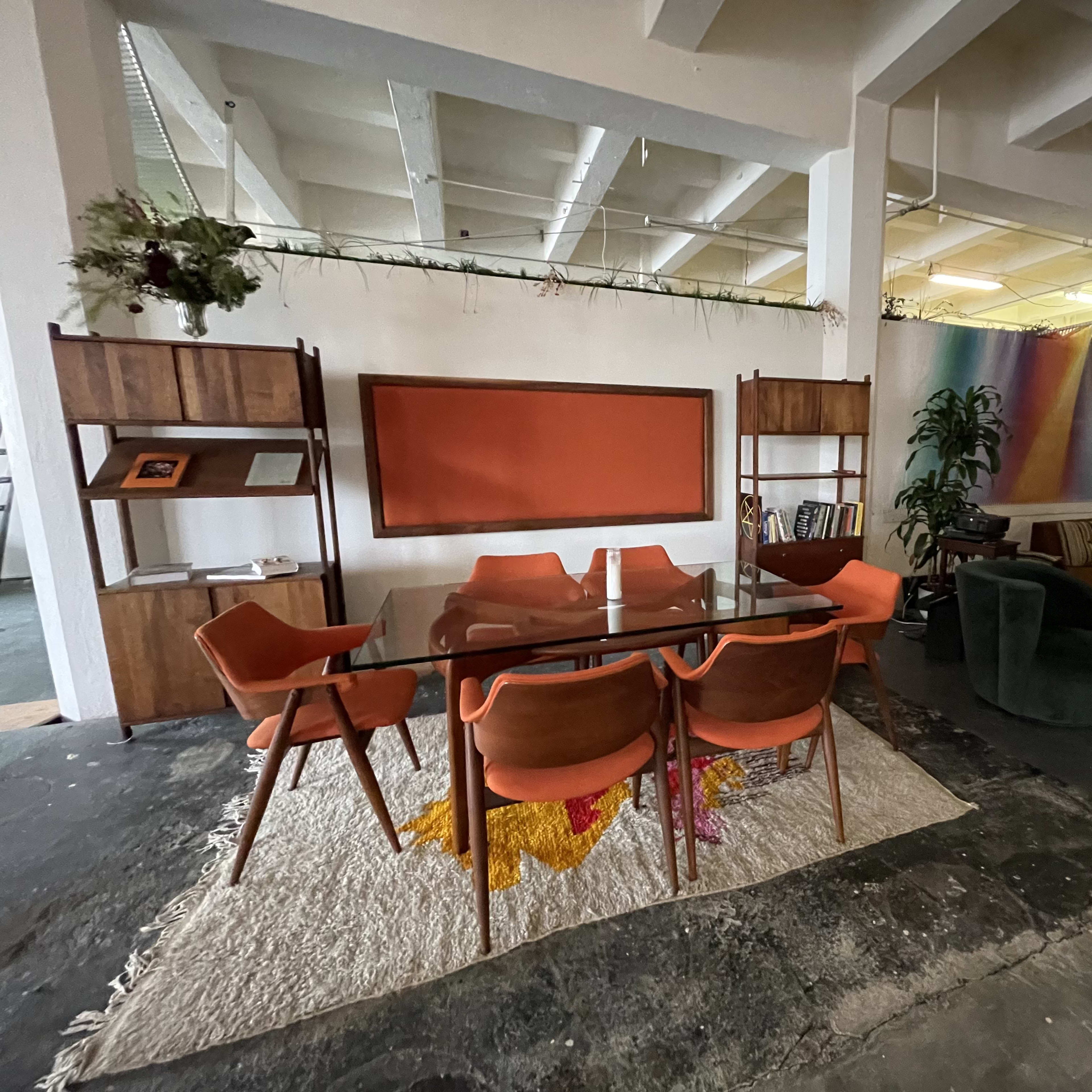 A modern open-plan office space featuring a glass dining table surrounded by orange chairs on a textured rug, a wooden bookshelf, and a cozy seating area with a green sofa and colorful artwork in the background.