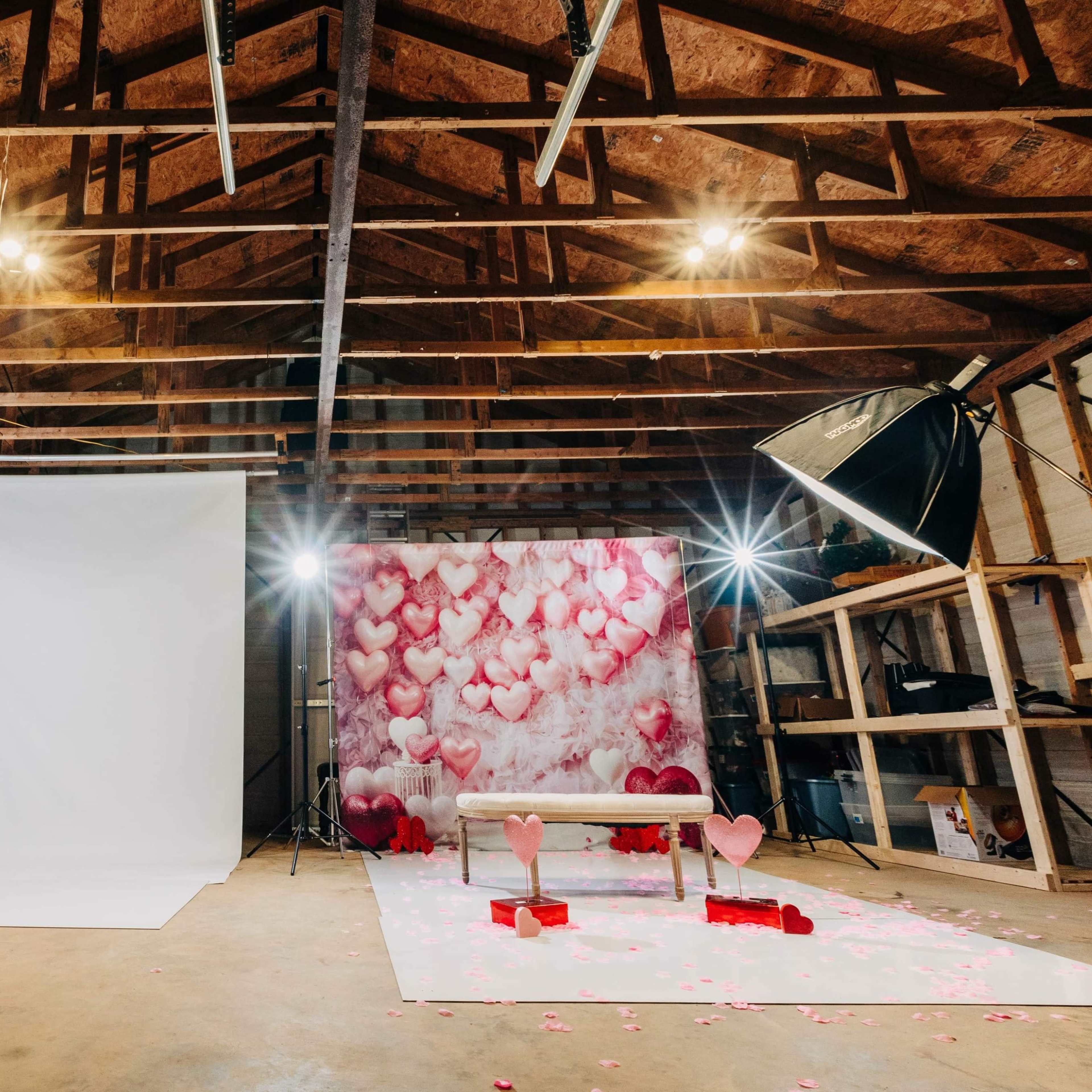A photography studio featuring a white backdrop, a pink heart-themed wall, and lighting equipment arranged around a bench with decorative hearts.