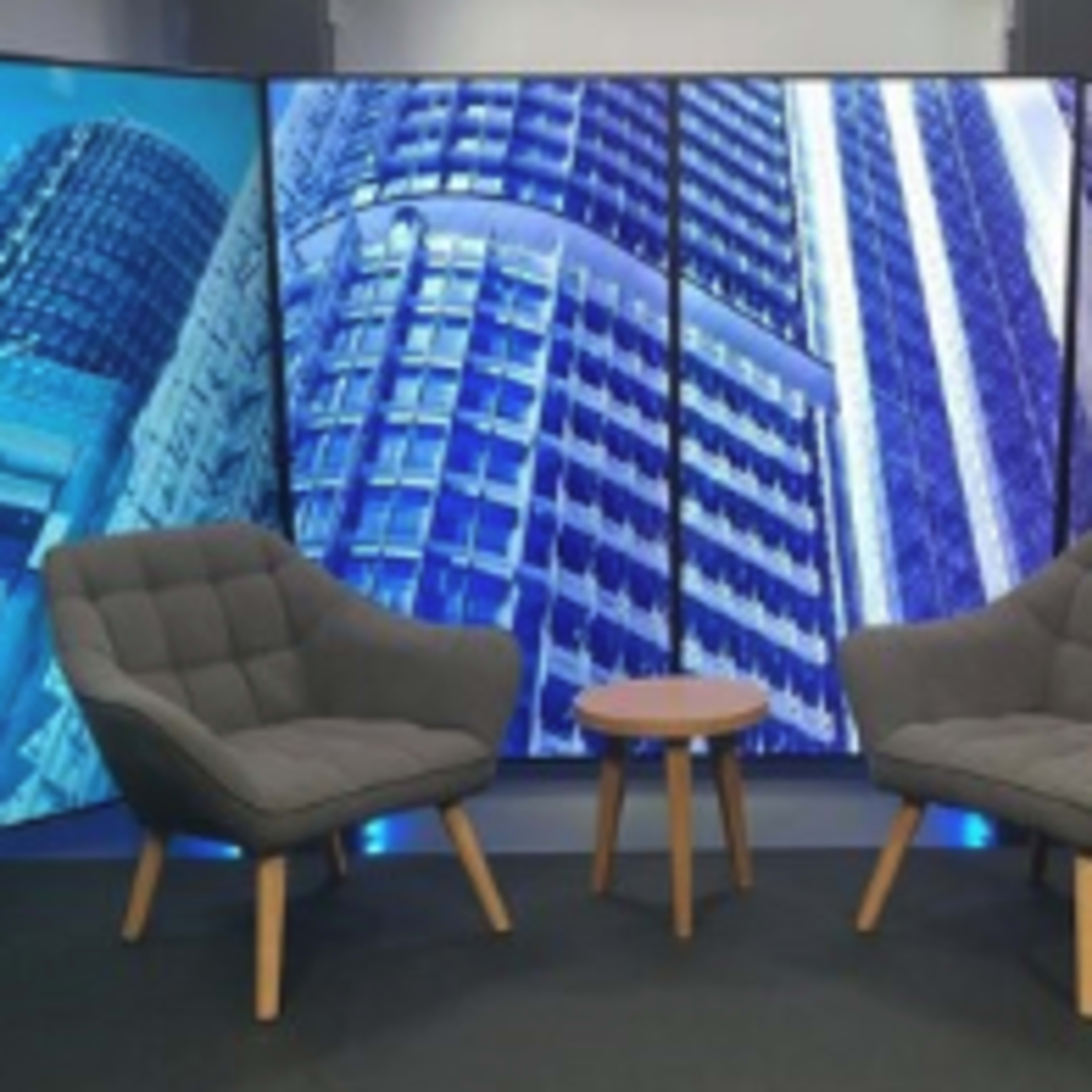 A modern studio set with two gray upholstered chairs facing a small wooden table, surrounded by backdrop panels featuring skyscraper images.