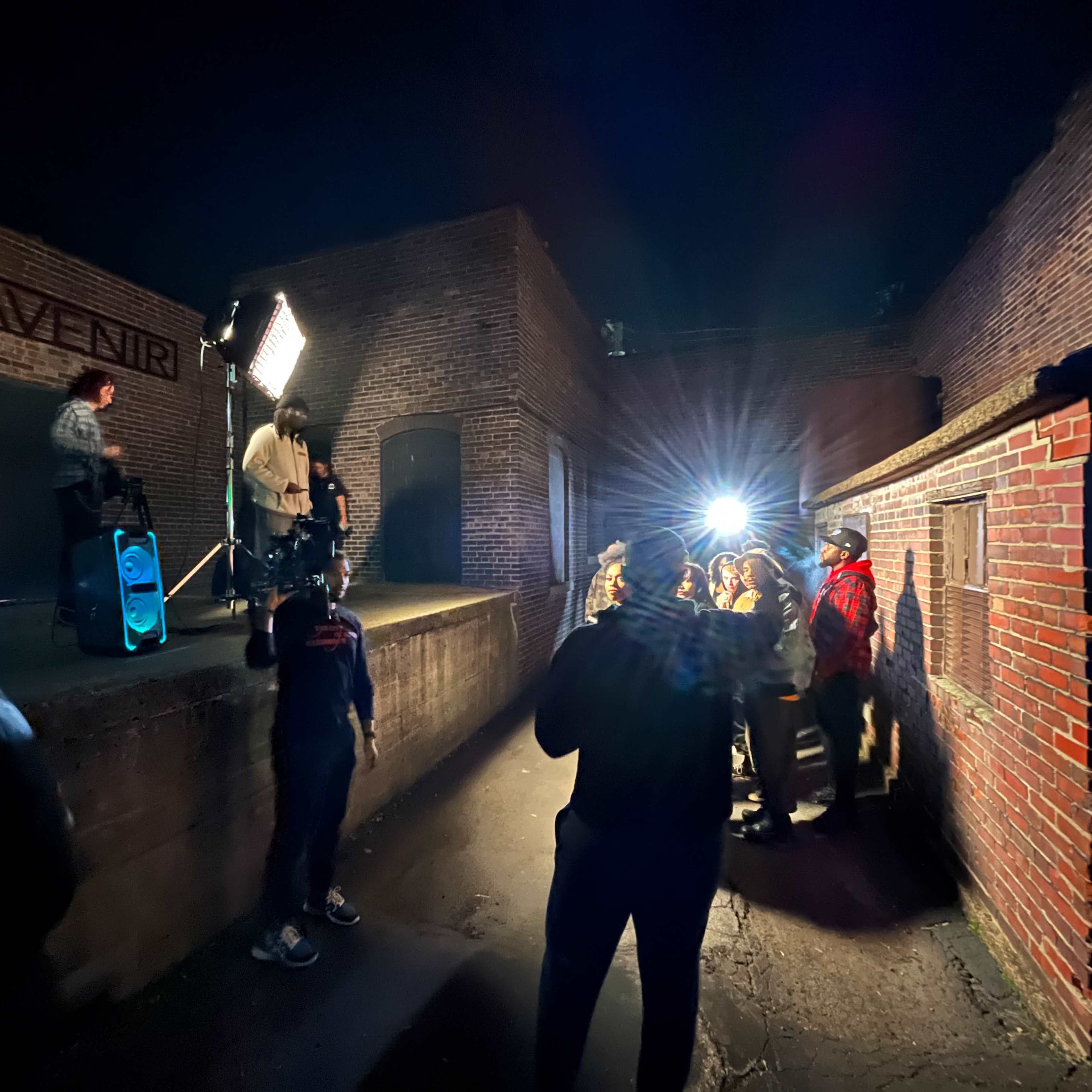 A group of people stands in a dimly lit outdoor area while a figure on a small platform speaks into a microphone, illuminated by bright lights.