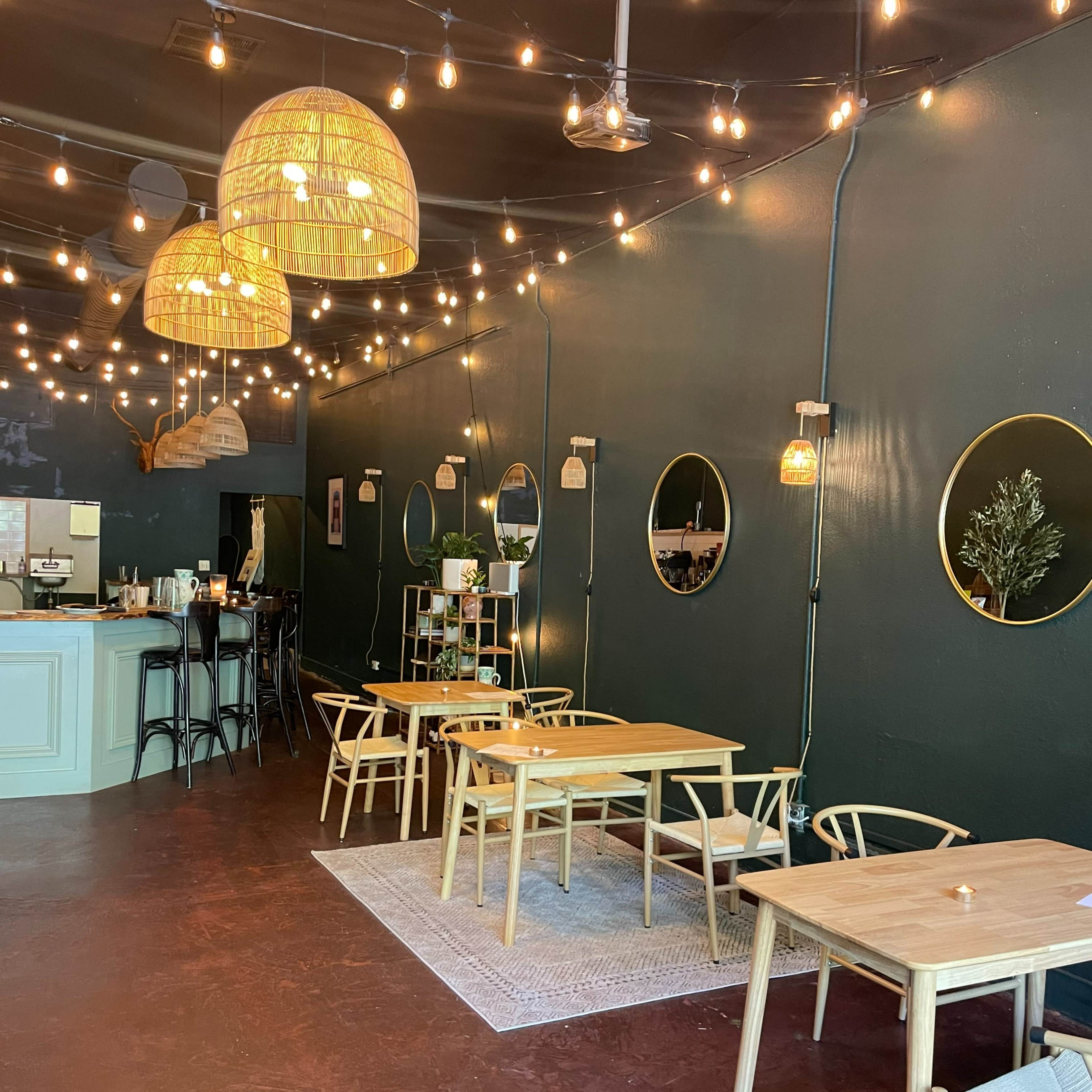 A cozy cafe interior with wooden tables, circular mirrors, and string lights hanging from a dark green wall.