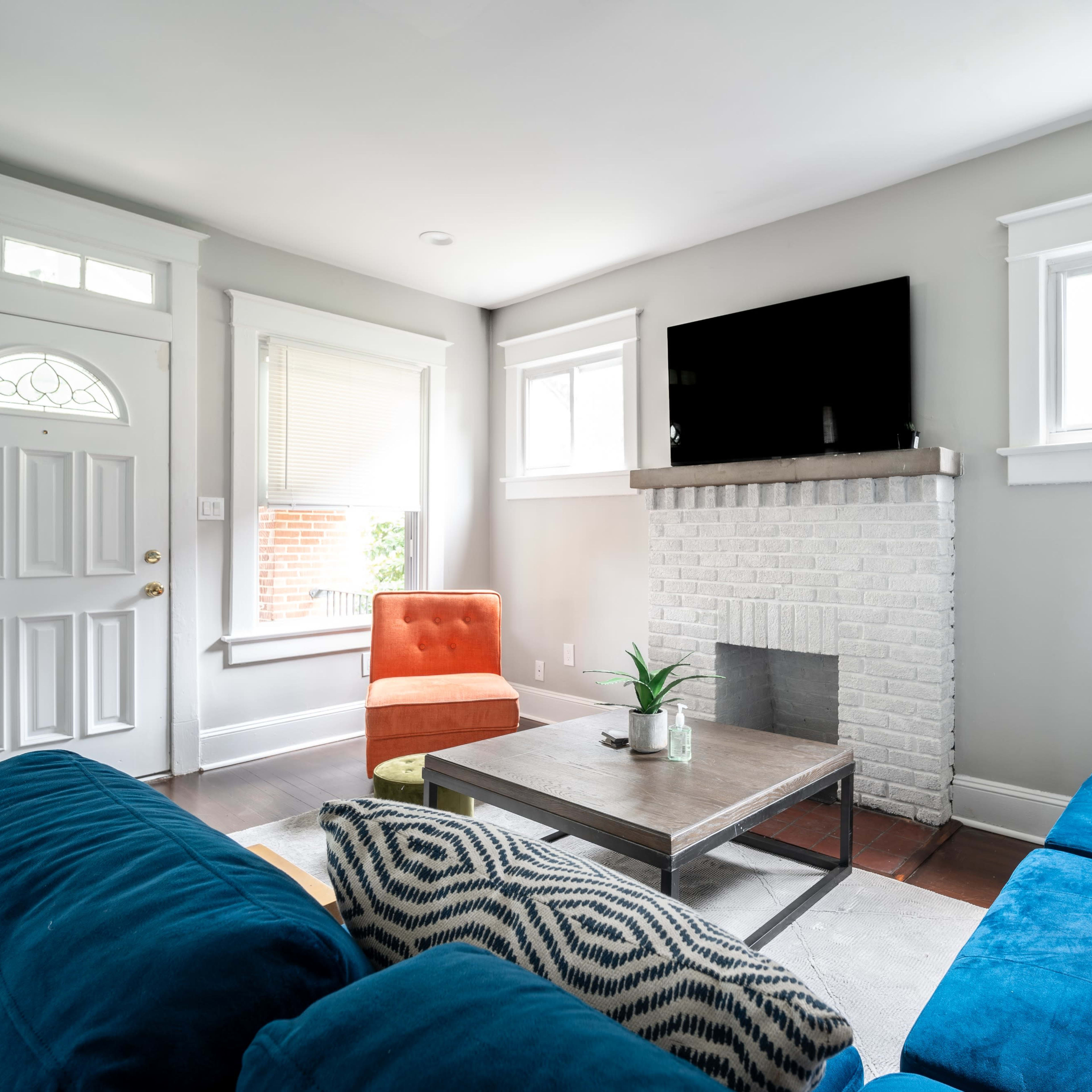A living room with a blue sofa, an orange armchair, a square coffee table, and a white brick fireplace, all under a mounted television.