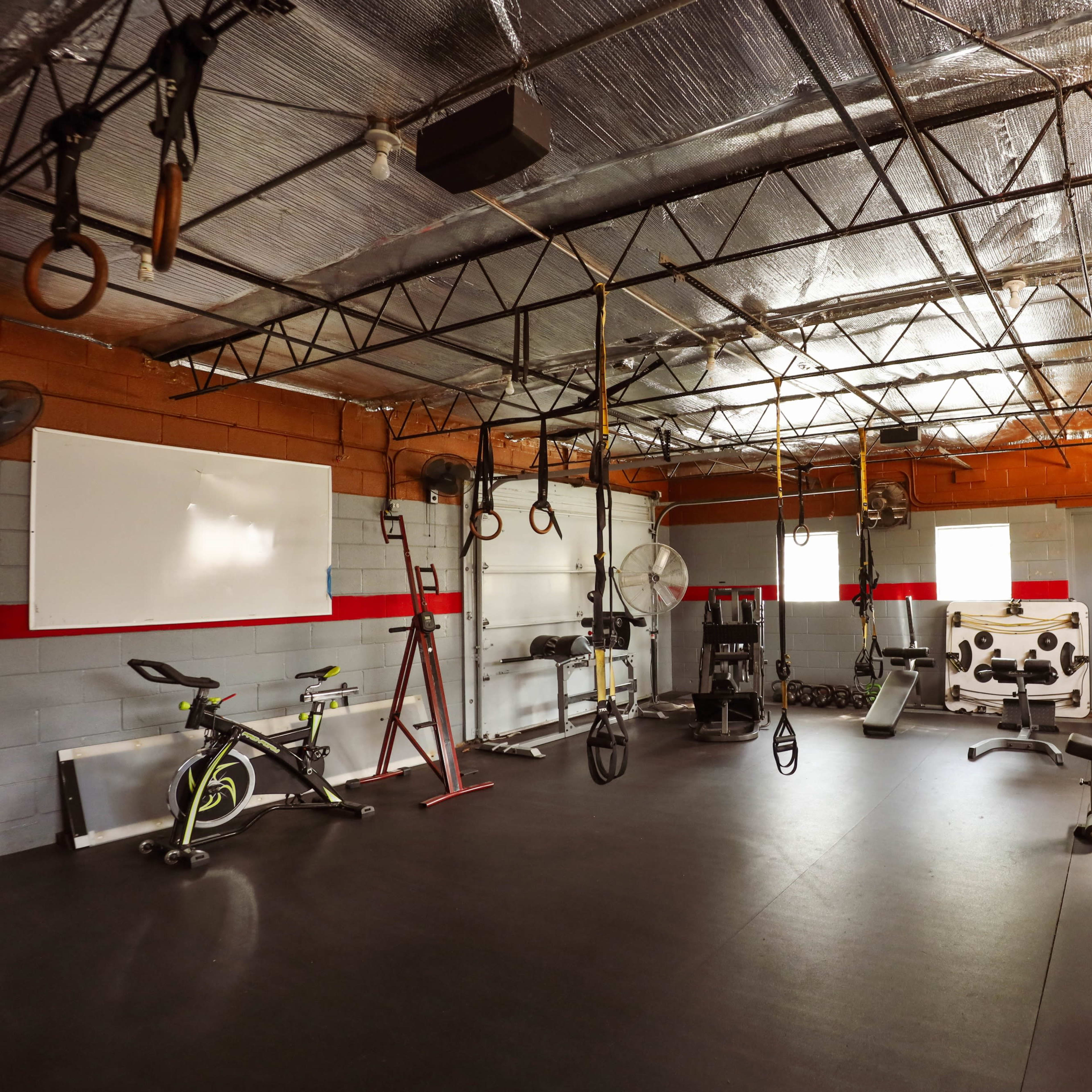 An empty gym with various exercise equipment, including a stationary bike, weight bench, and hanging straps, set against bare walls and exposed beams.