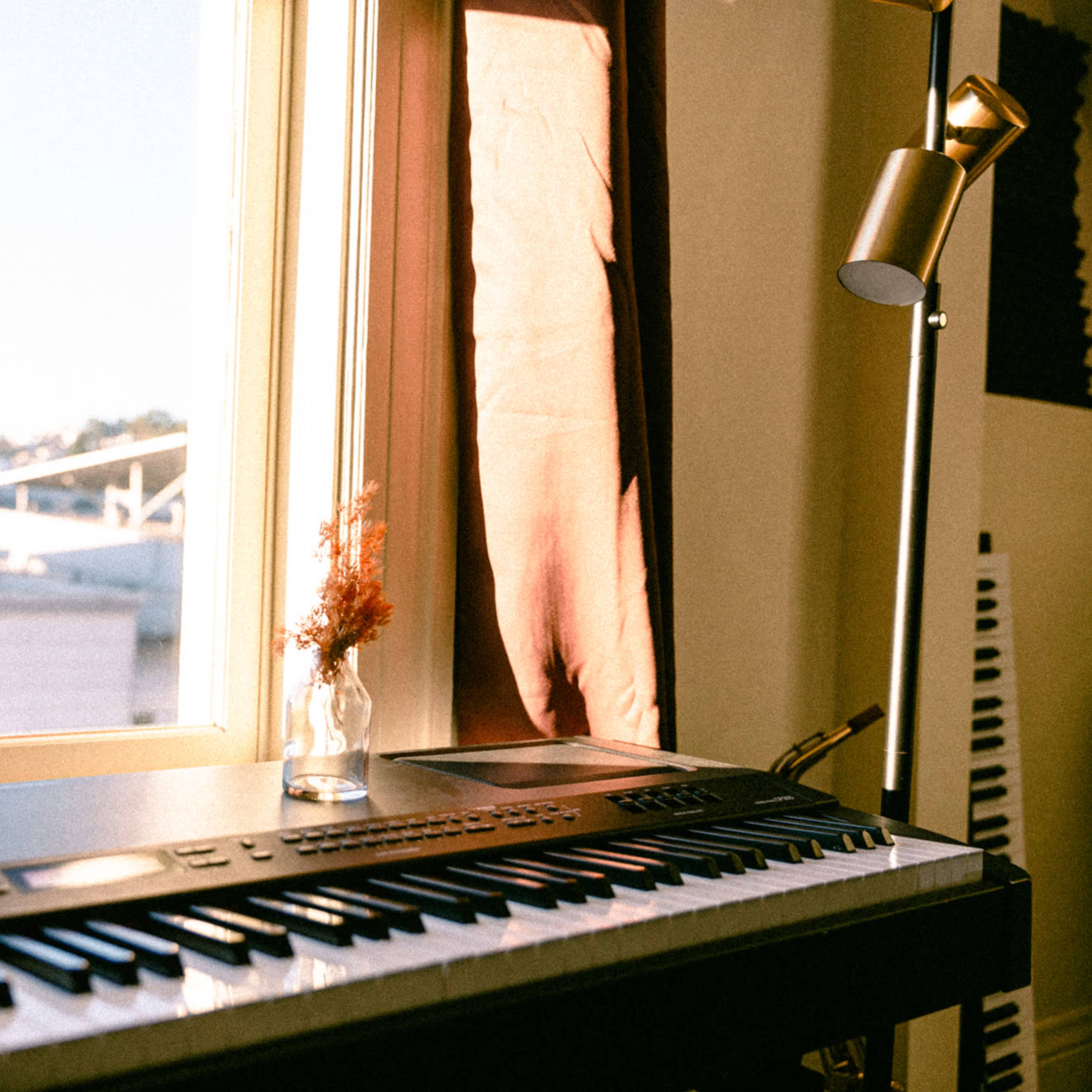 A keyboard sits next to a window, with a small vase of dried flowers in front and a lamp illuminating the scene.