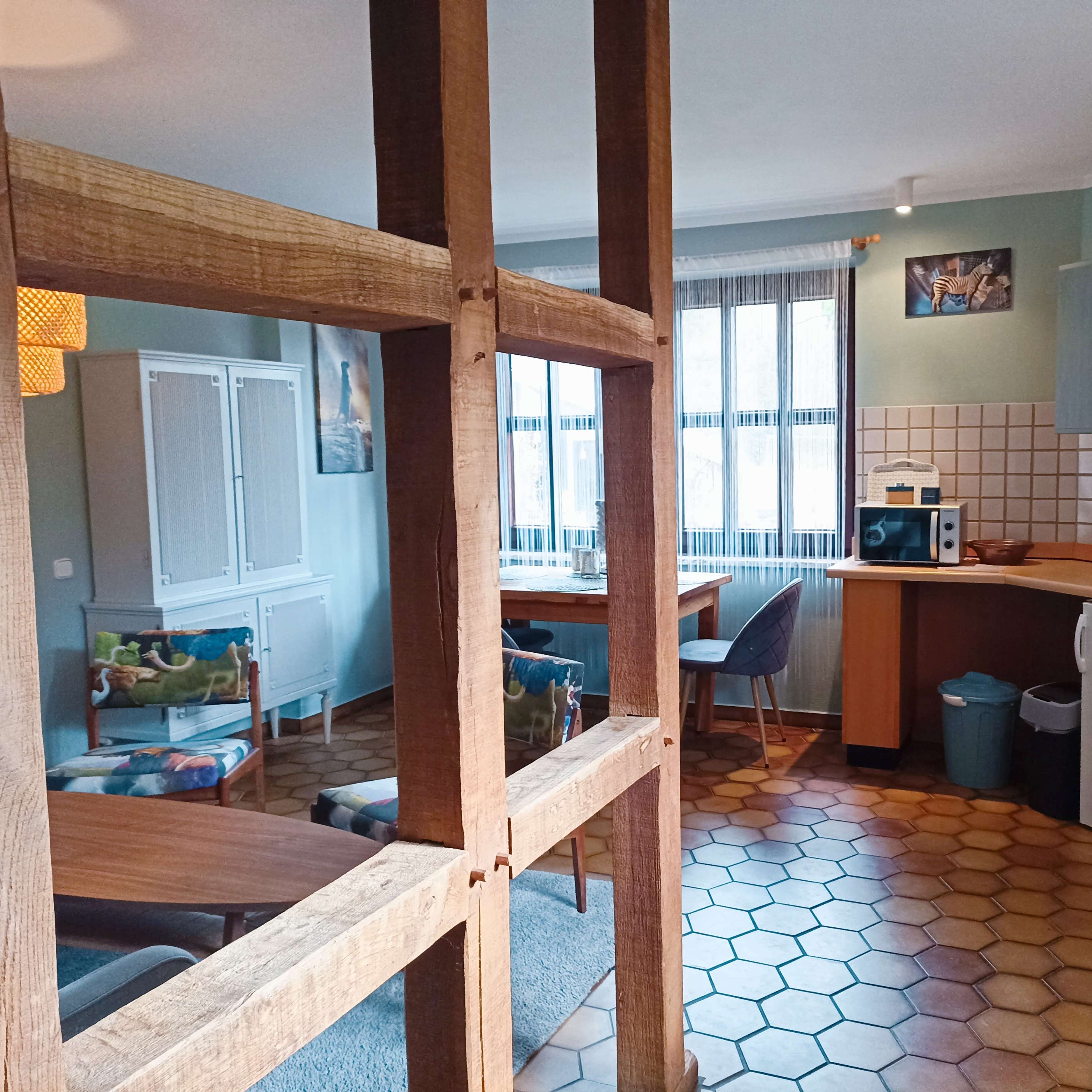 A cozy kitchen and living area separated by a wooden frame, with a dining table, colorful chairs, and appliances visible.
