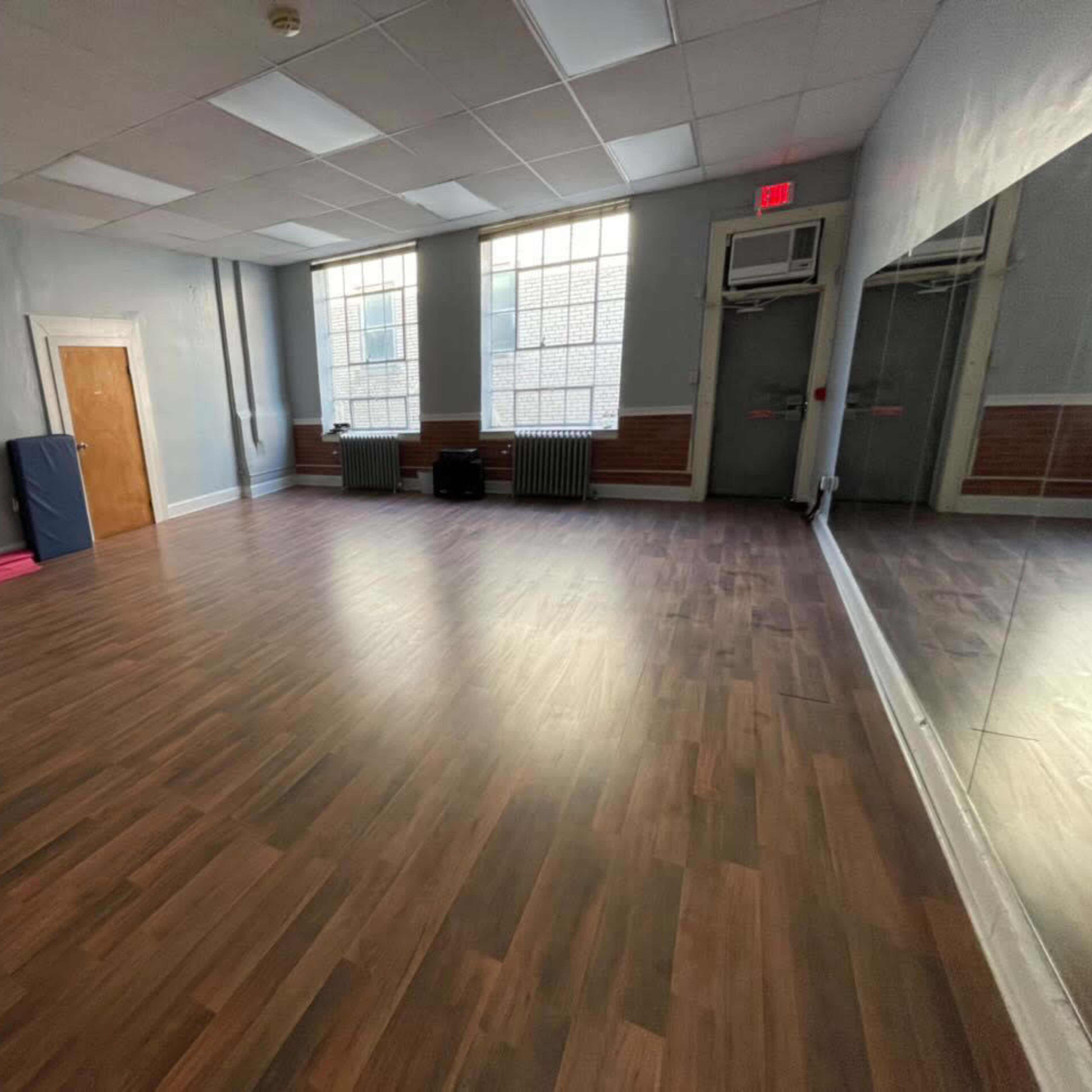 A vacant room with wooden flooring, large windows, and a mirrored wall, furnished with a few stacked chairs and exercise mats.