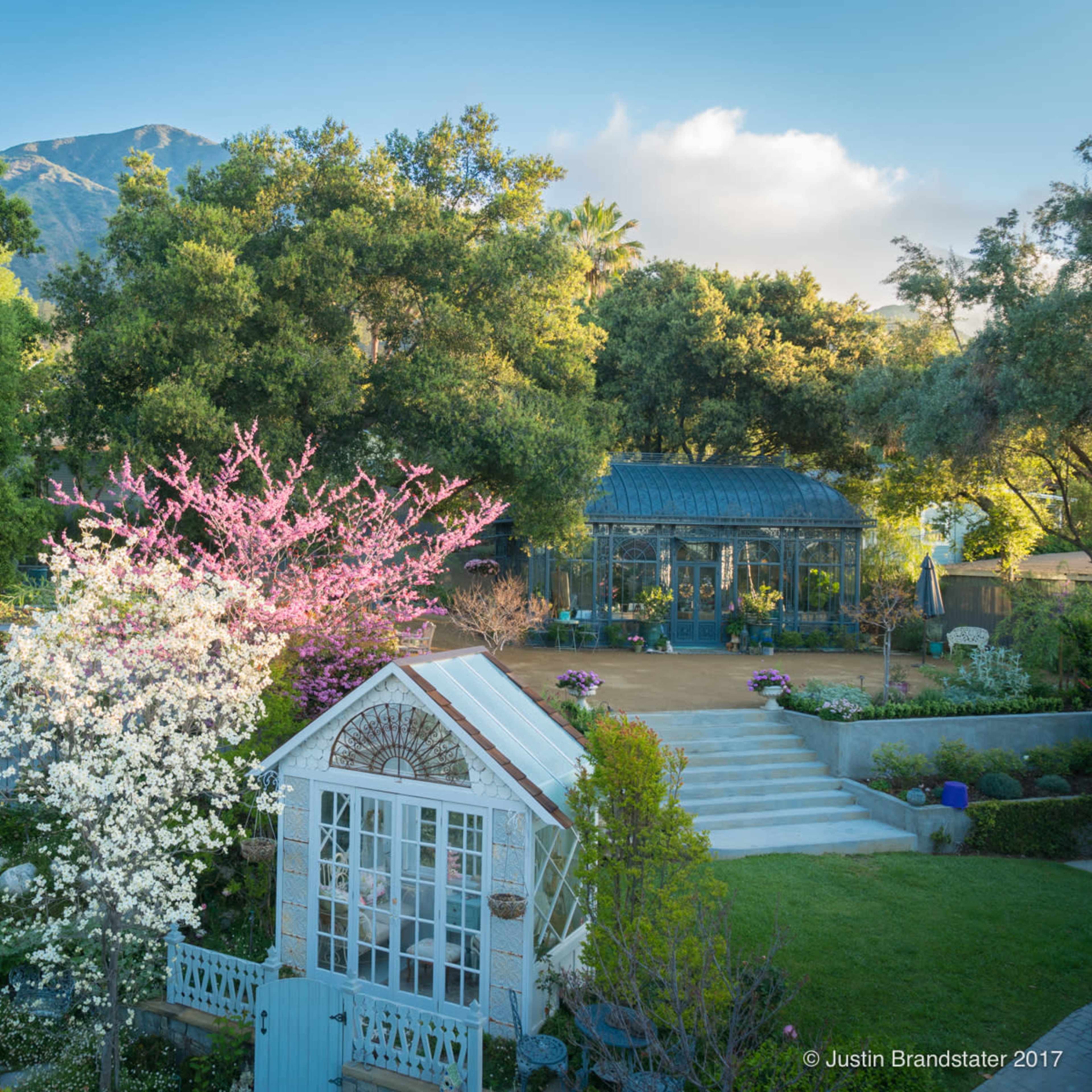 A landscaped garden featuring a white gazebo, blooming trees, and a greenhouse surrounded by lush greenery and pathways.