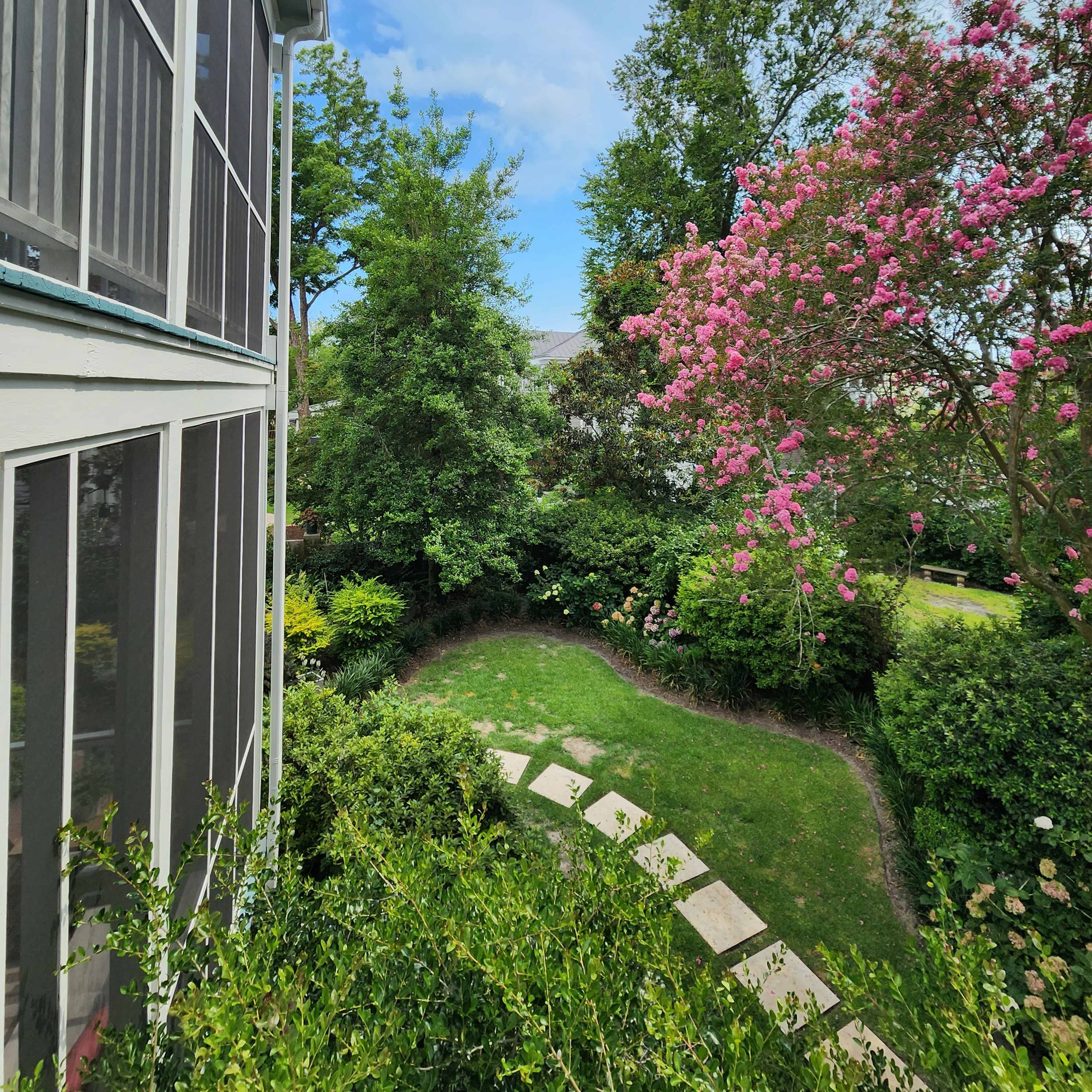 A pathway of stone steps winds through a lush garden with trees and flowering bushes beside a screened porch.