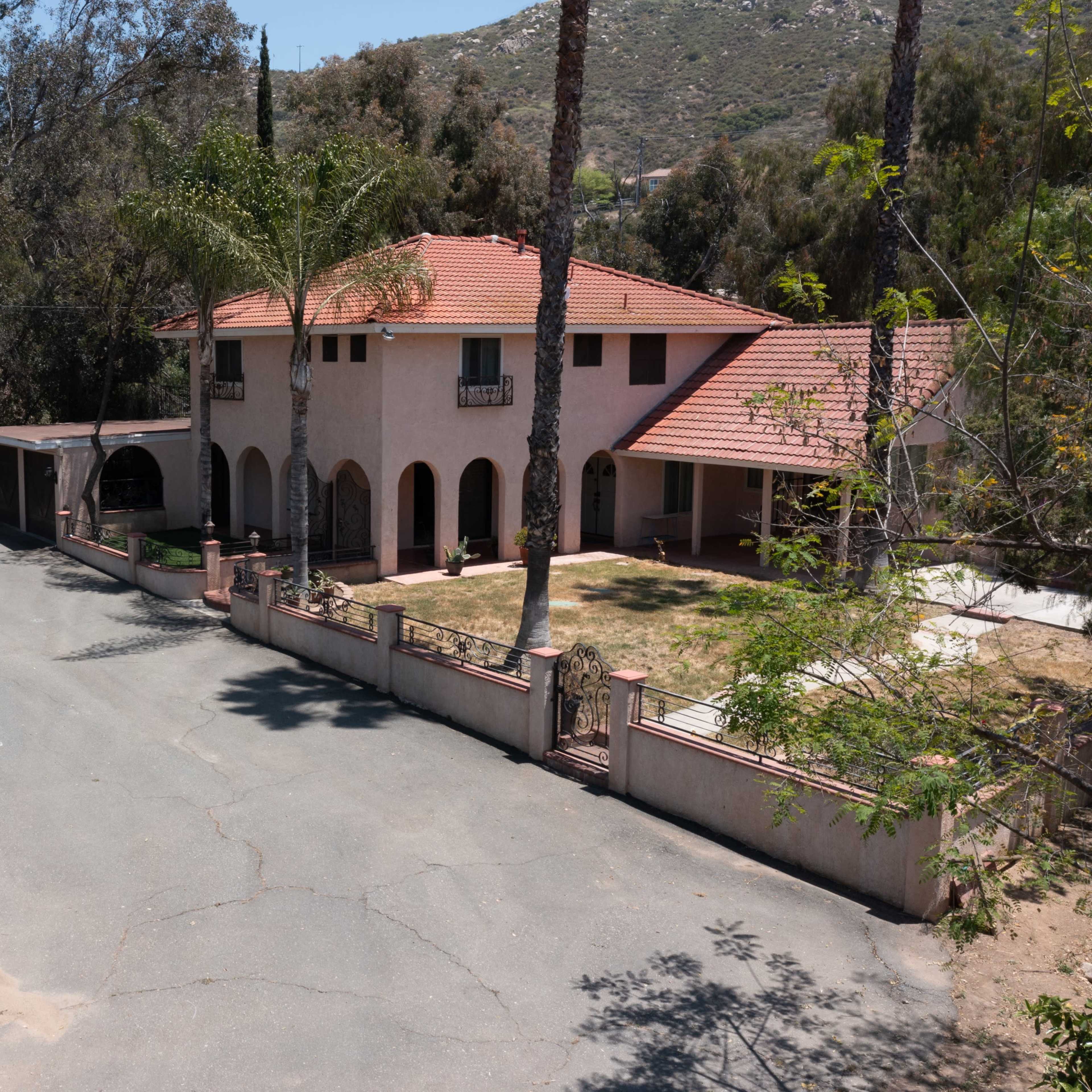 A large two-story house with a red tiled roof is situated at the end of a winding, tree-lined street, with a palm tree and a green lawn in the front yard.