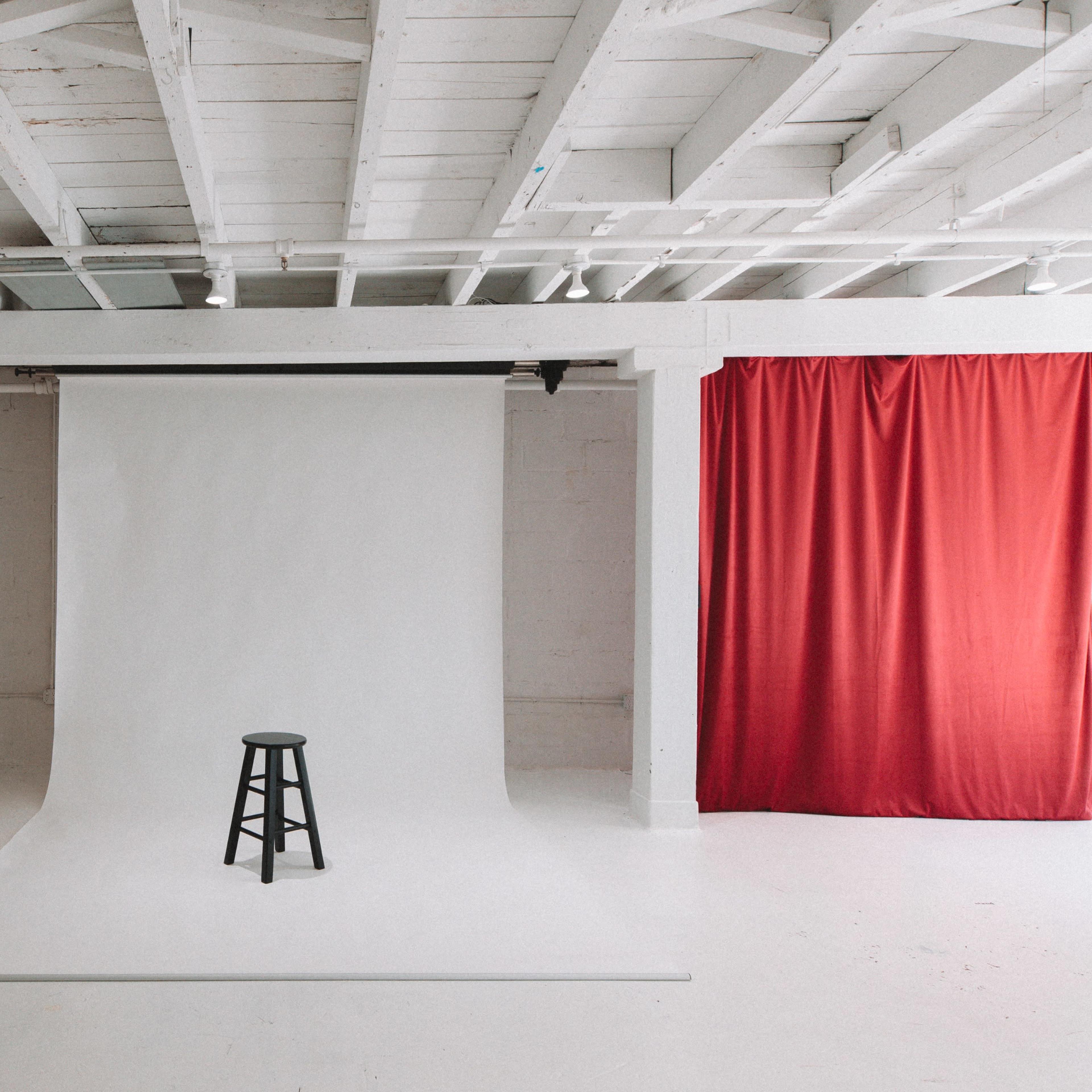 A photography studio with a white backdrop, a black stool in the center, and red drapery on one side.