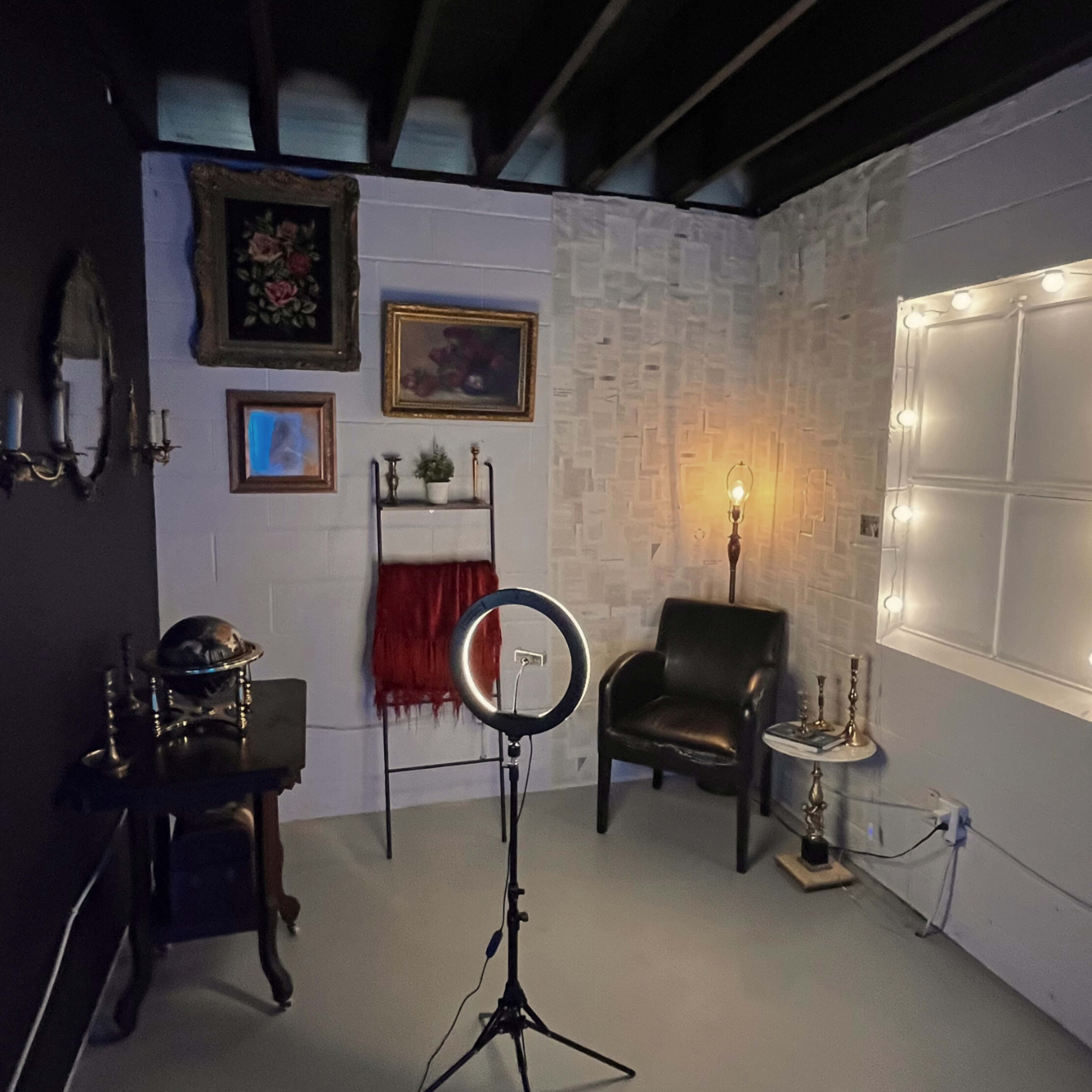 A small, dimly lit room featuring a black wall, assorted artwork, a ring light on a stand, a chair, and decorative items on a shelf.