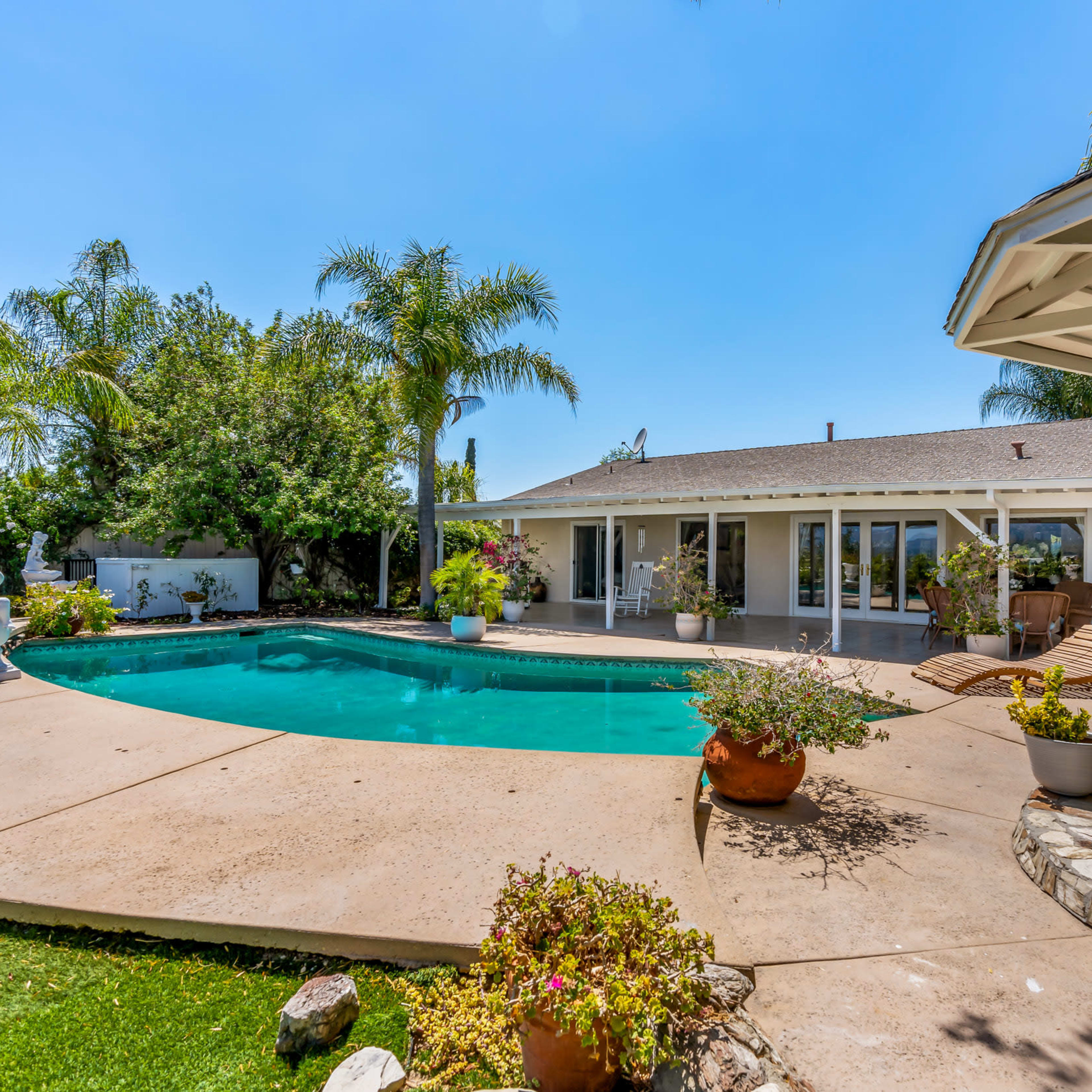 A backyard with a clear swimming pool, surrounded by greenery and a patio with seating areas.