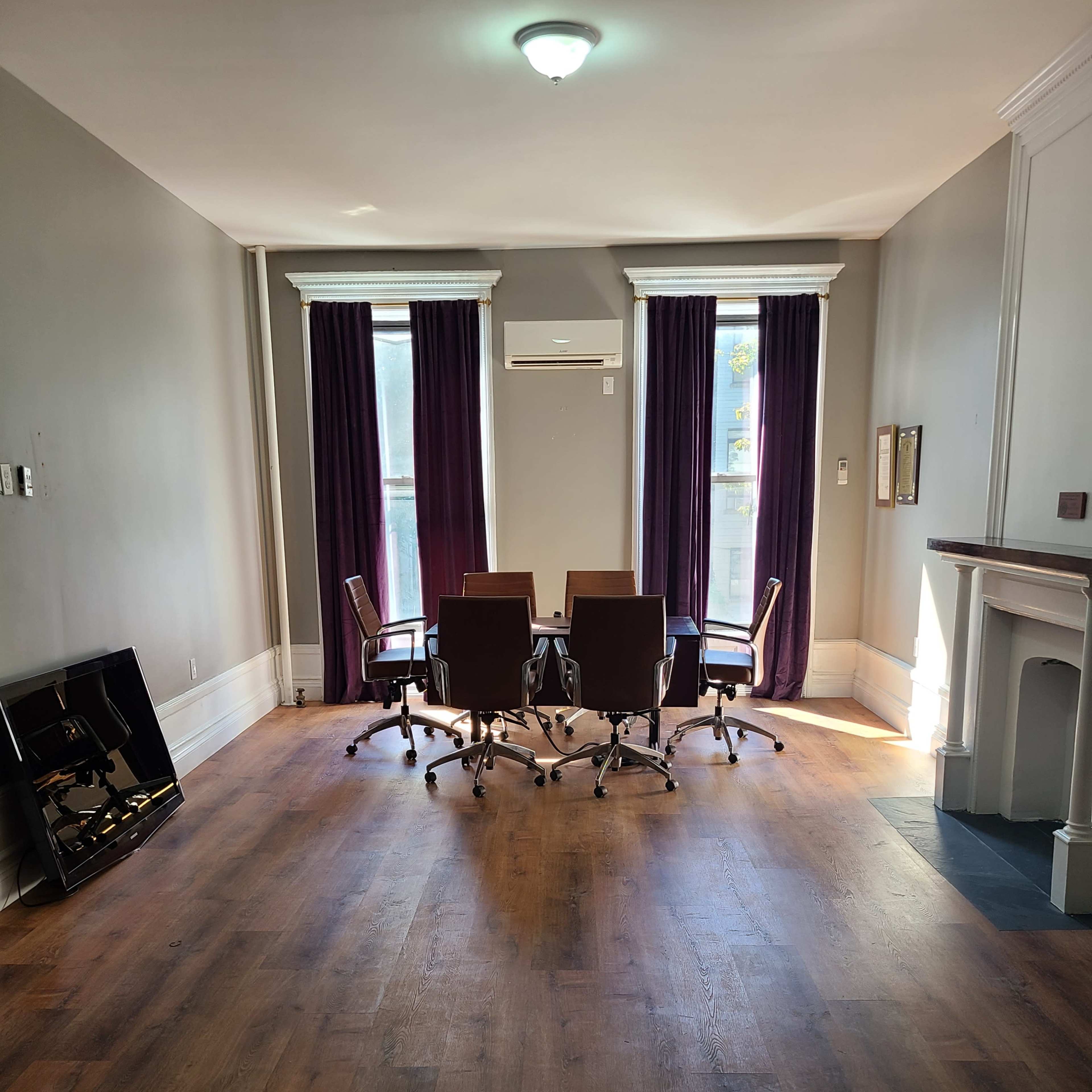 A spacious meeting room features a circular arrangement of five swivel chairs around a central table, with large windows dressed in purple curtains.