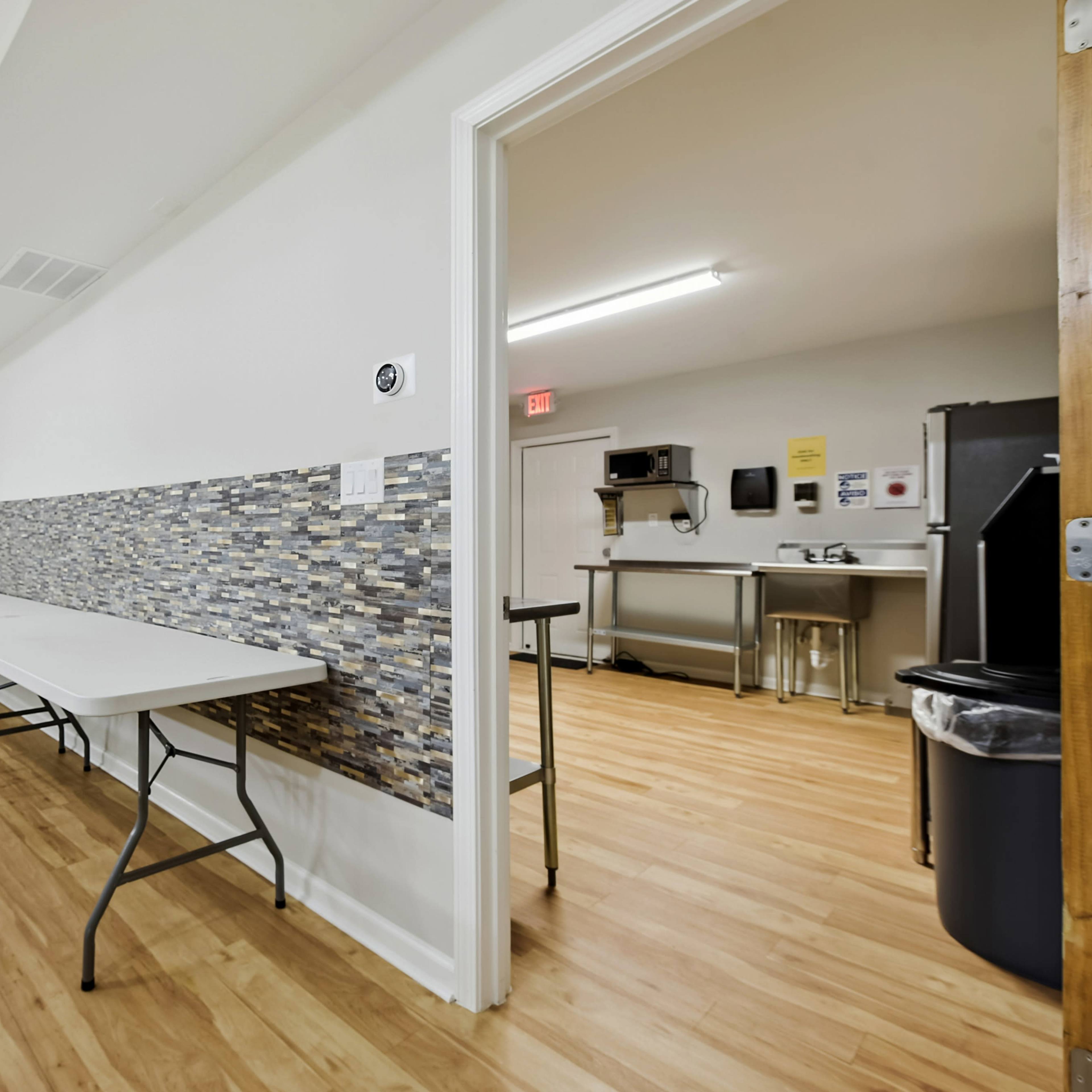 A hallway with light-colored wooden flooring, leading to a kitchen area, and features two folding tables against a textured wall.