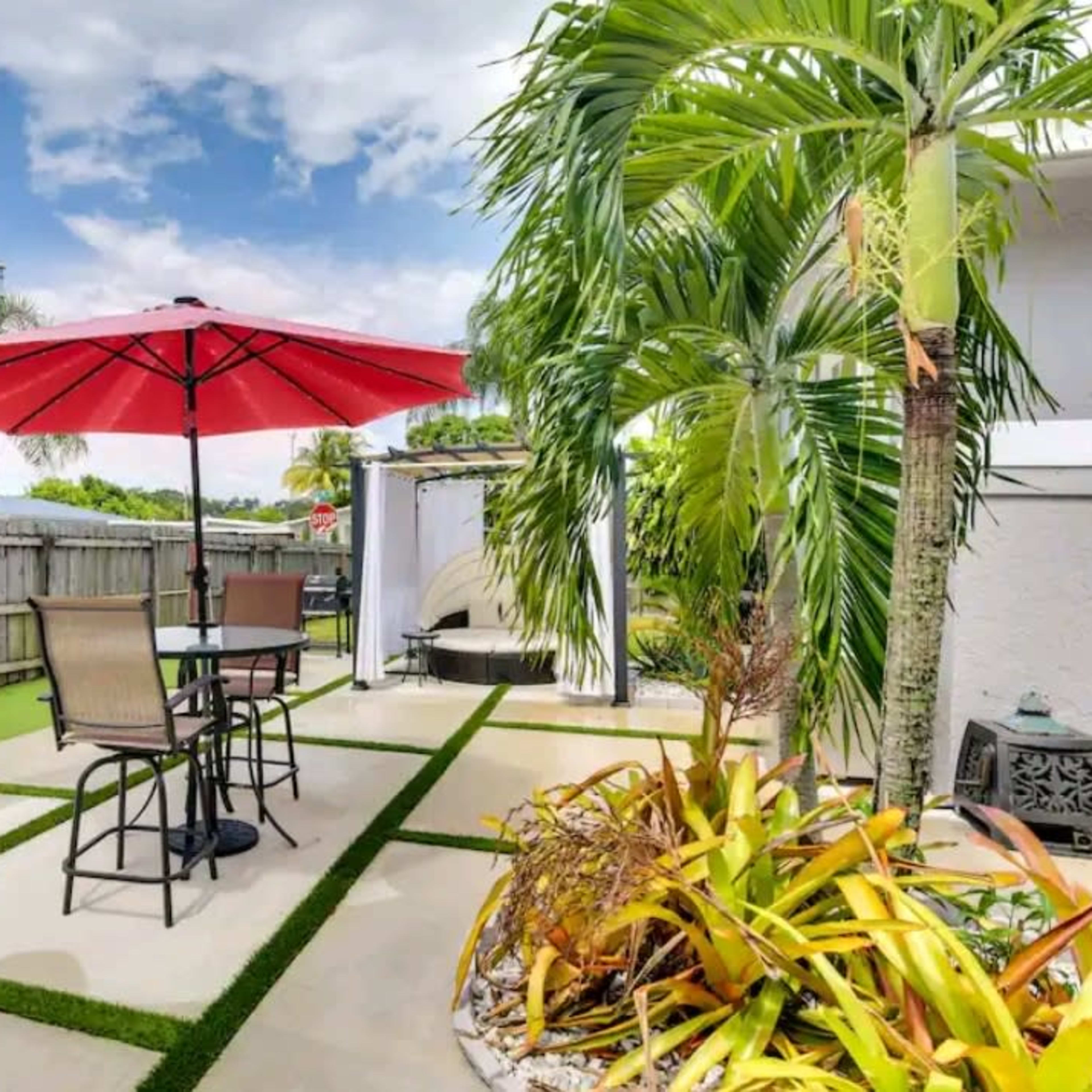 A backyard patio featuring a red umbrella over a table with chairs, surrounded by landscaping and a grill area.