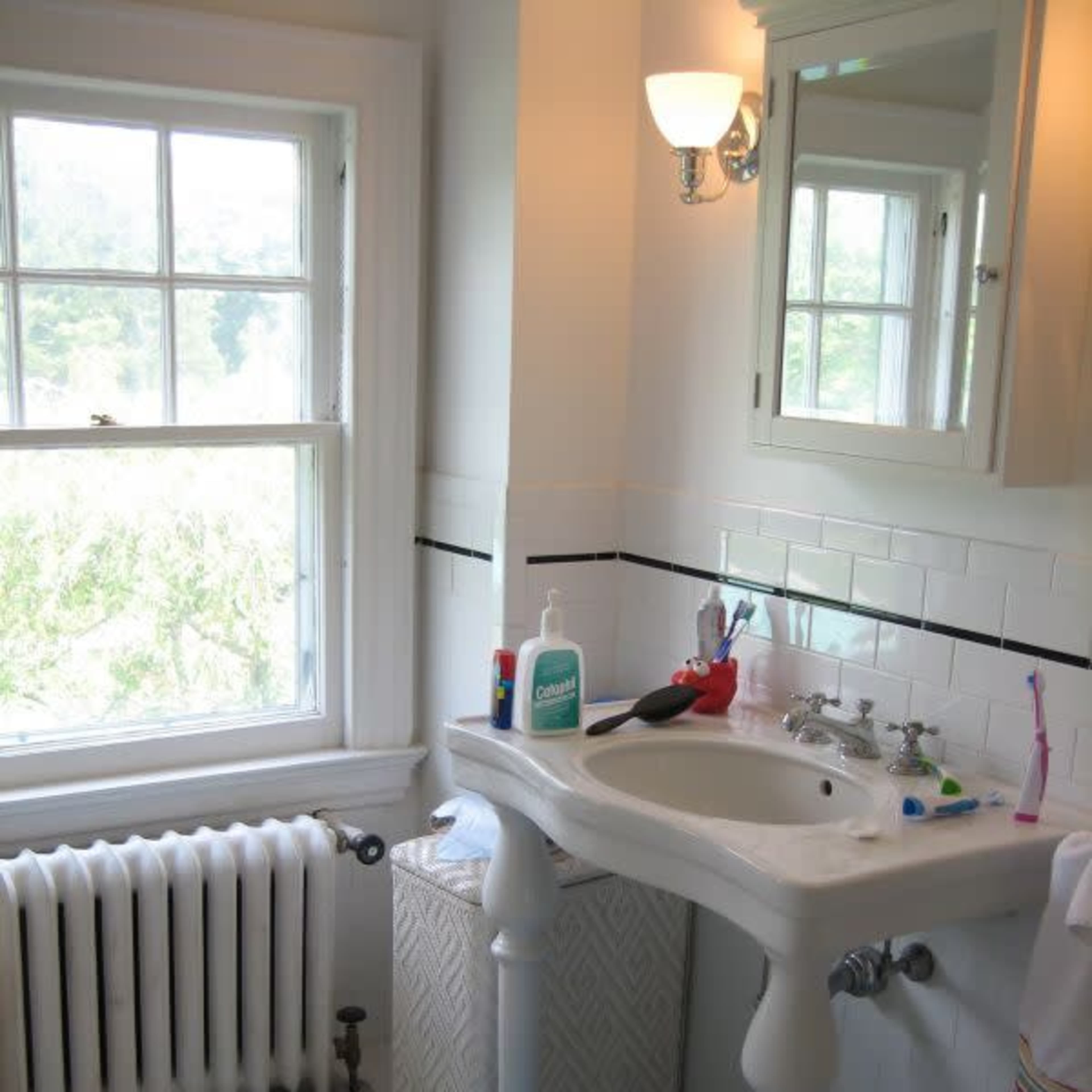 The bathroom features a white pedestal sink, a mirror above it, a towel hanging on a rack, and a blue toy stool next to a patterned wastebasket.
