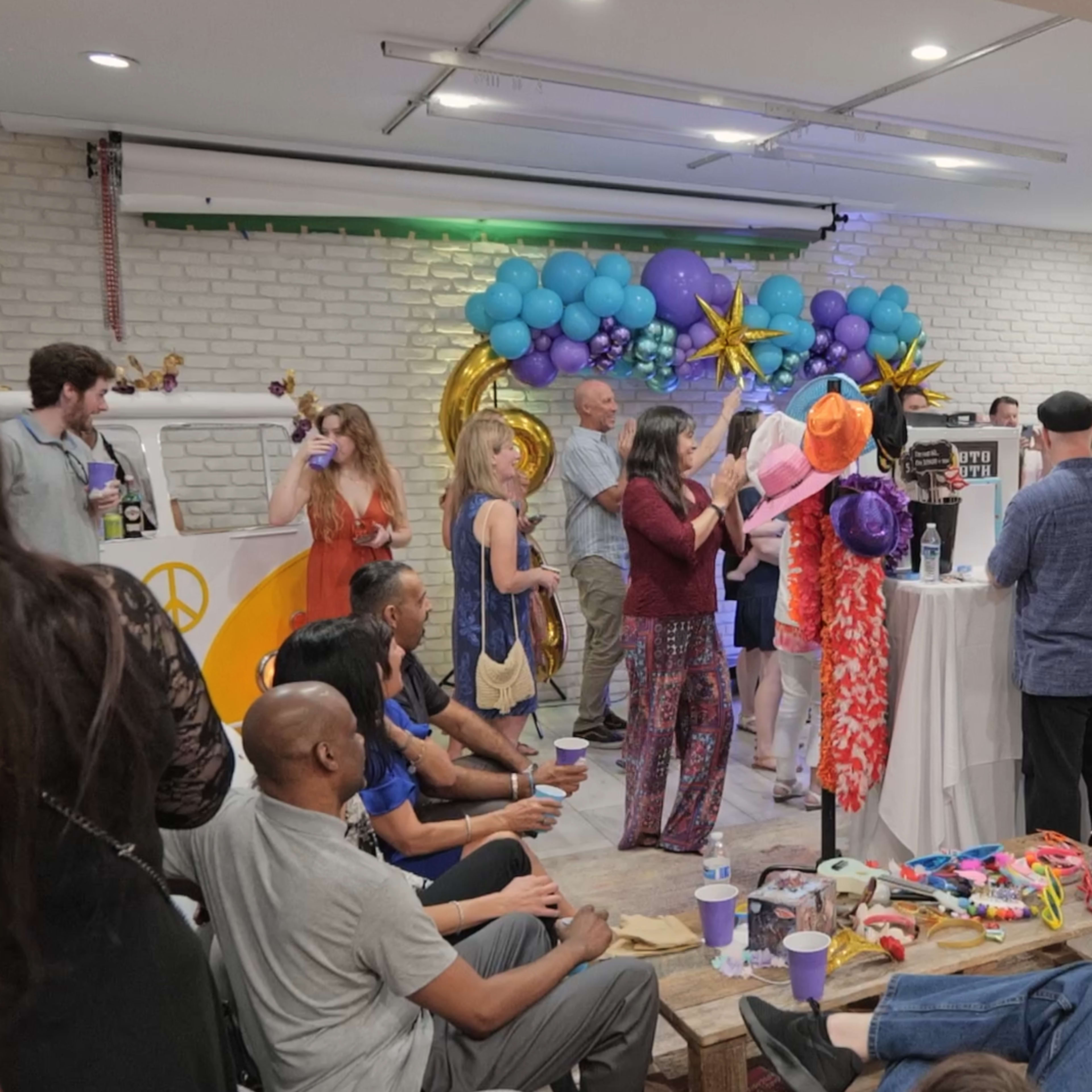 A group of people gathers in a brightly decorated room, socializing around a table filled with food and colorful party supplies.