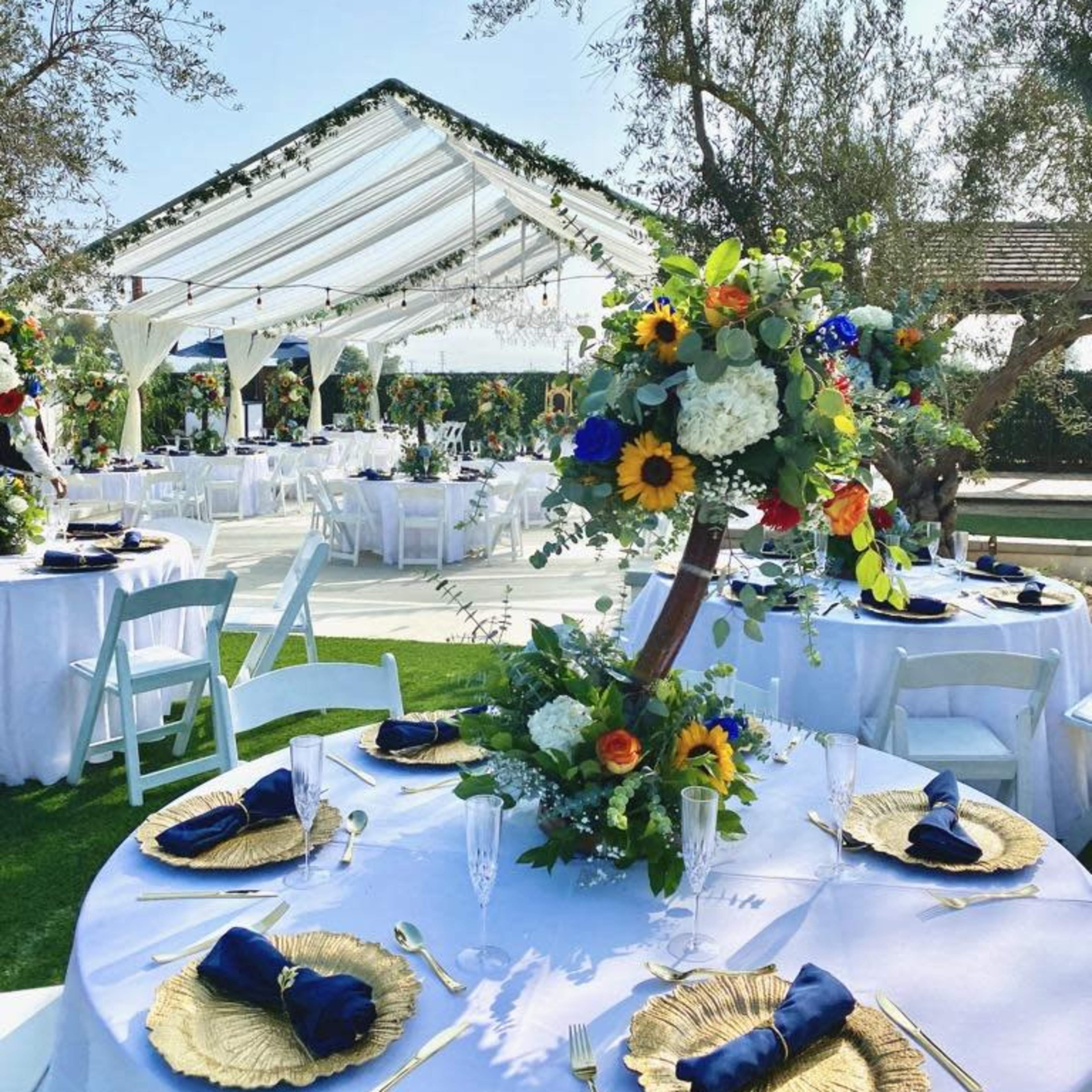 A decorated outdoor wedding venue features round tables set with gold charger plates, navy napkins, and vibrant floral centerpieces under a white canopy.