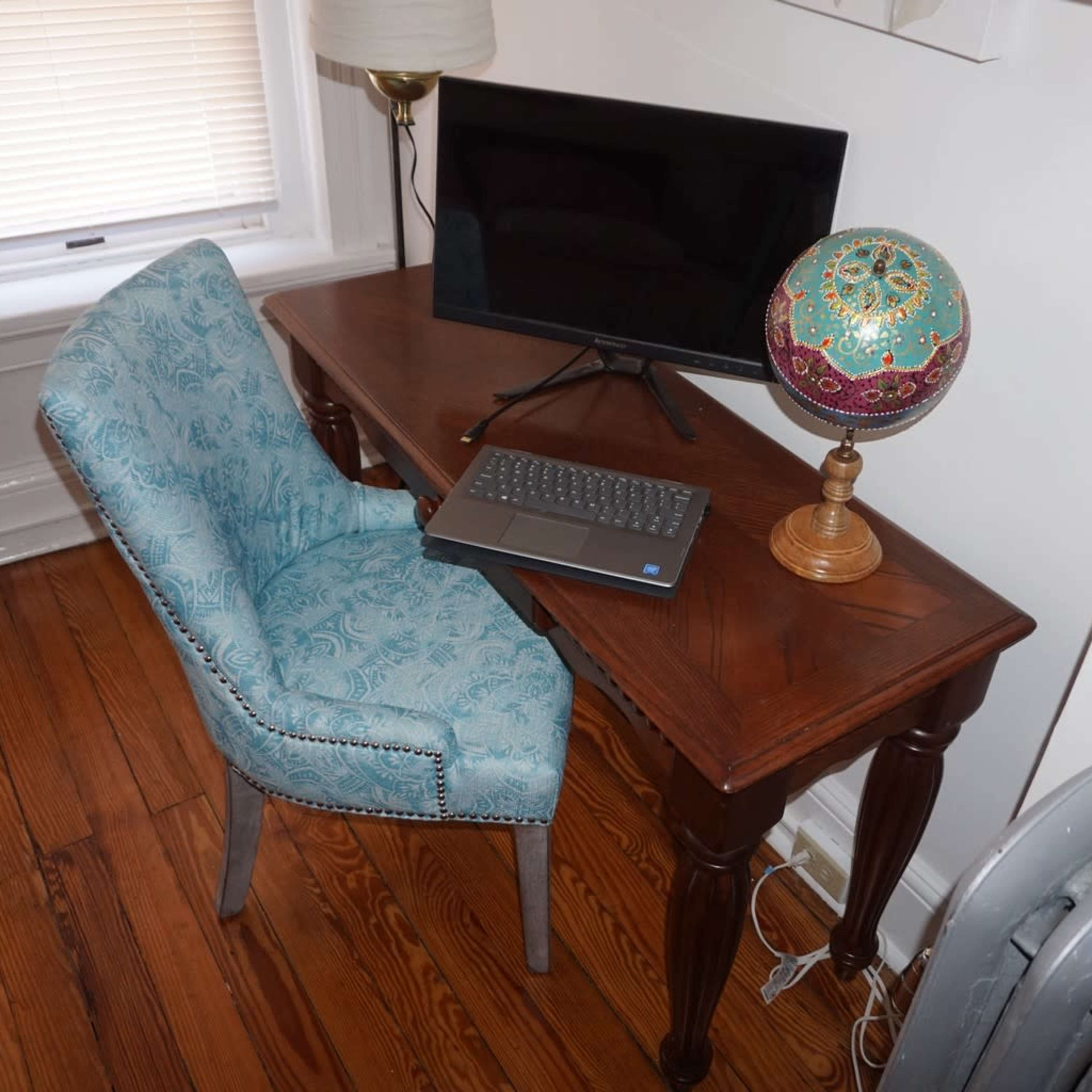 A wooden desk with a computer monitor, a laptop, a decorative globe, and a blue upholstered chair sits on a wooden floor.