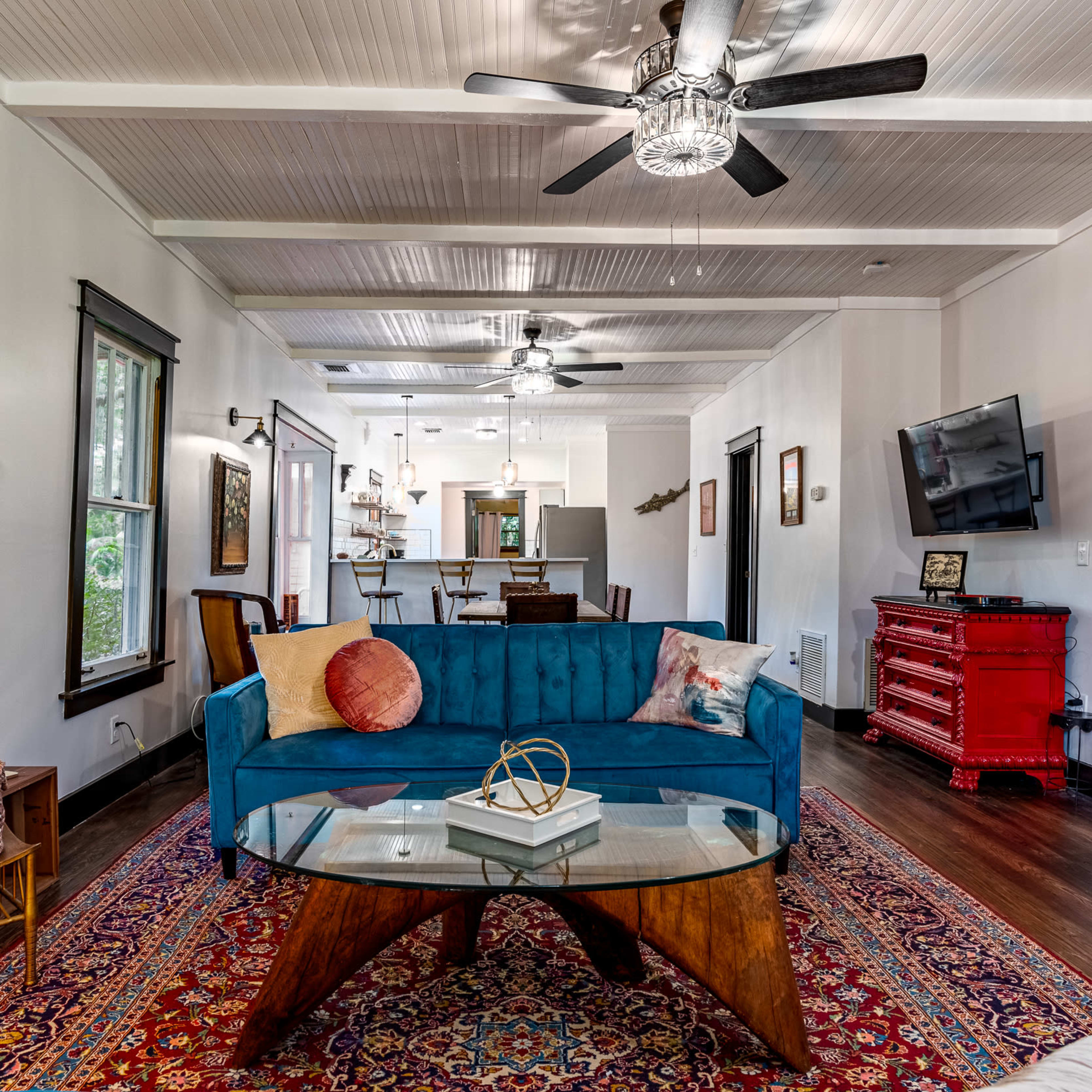 A living room featuring a blue velvet sofa, a glass-top coffee table, and a patterned rug, with an open kitchen visible in the background.