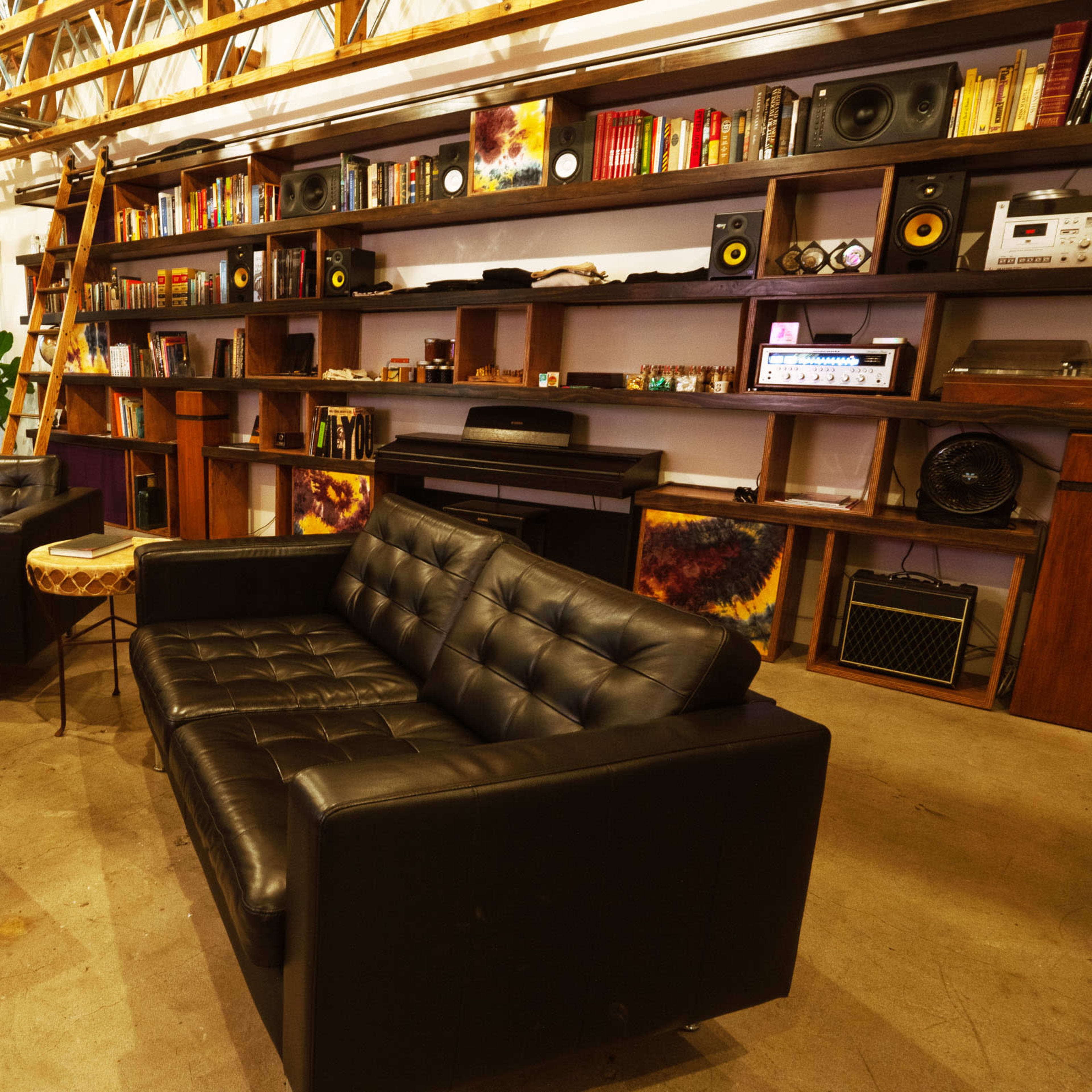A cozy interior space with black leather sofas, a wooden coffee table, and a bookshelf filled with various items, illuminated by overhead lights.