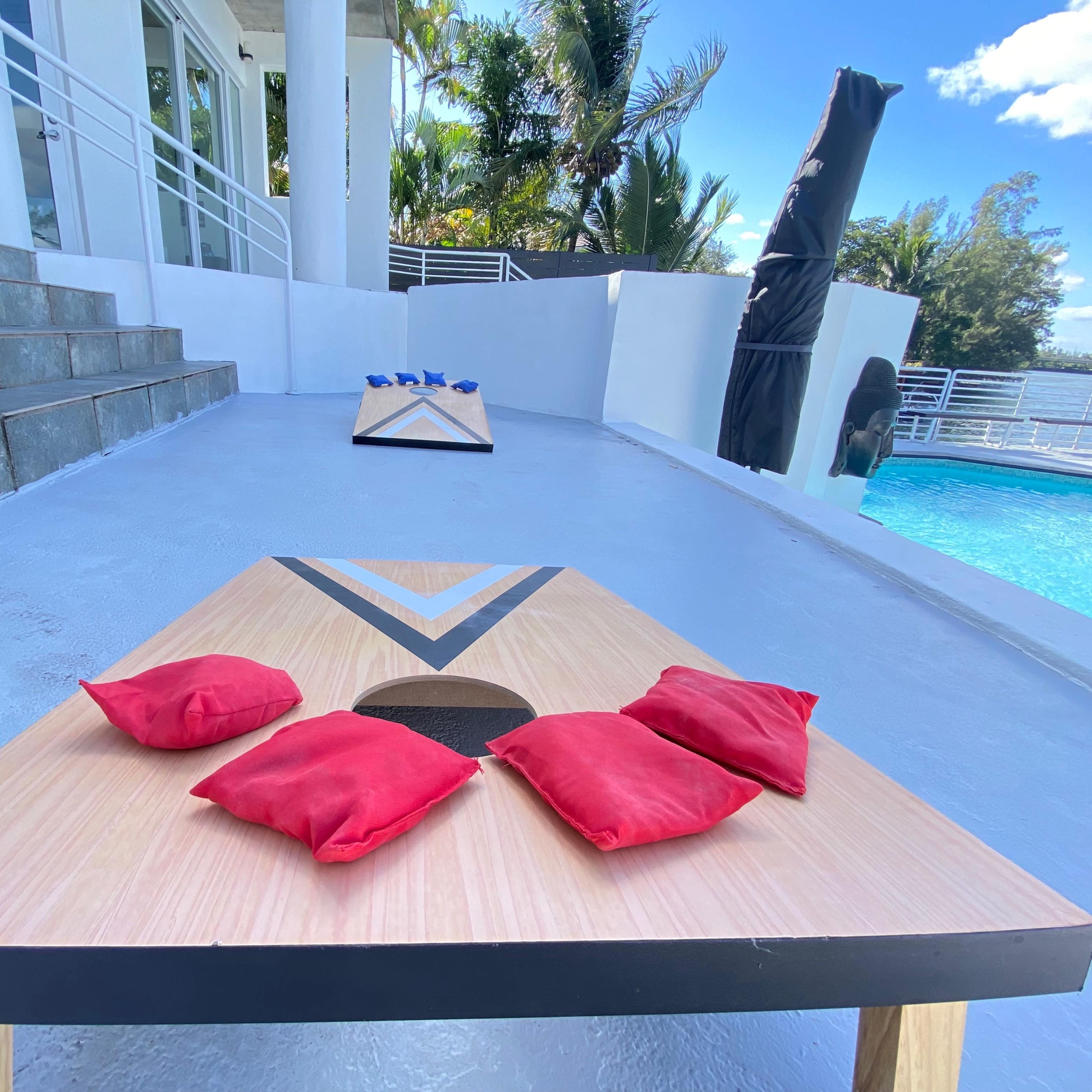 A set of cornhole boards with red bean bags placed in front of them, situated near a poolside area surrounded by palm trees.