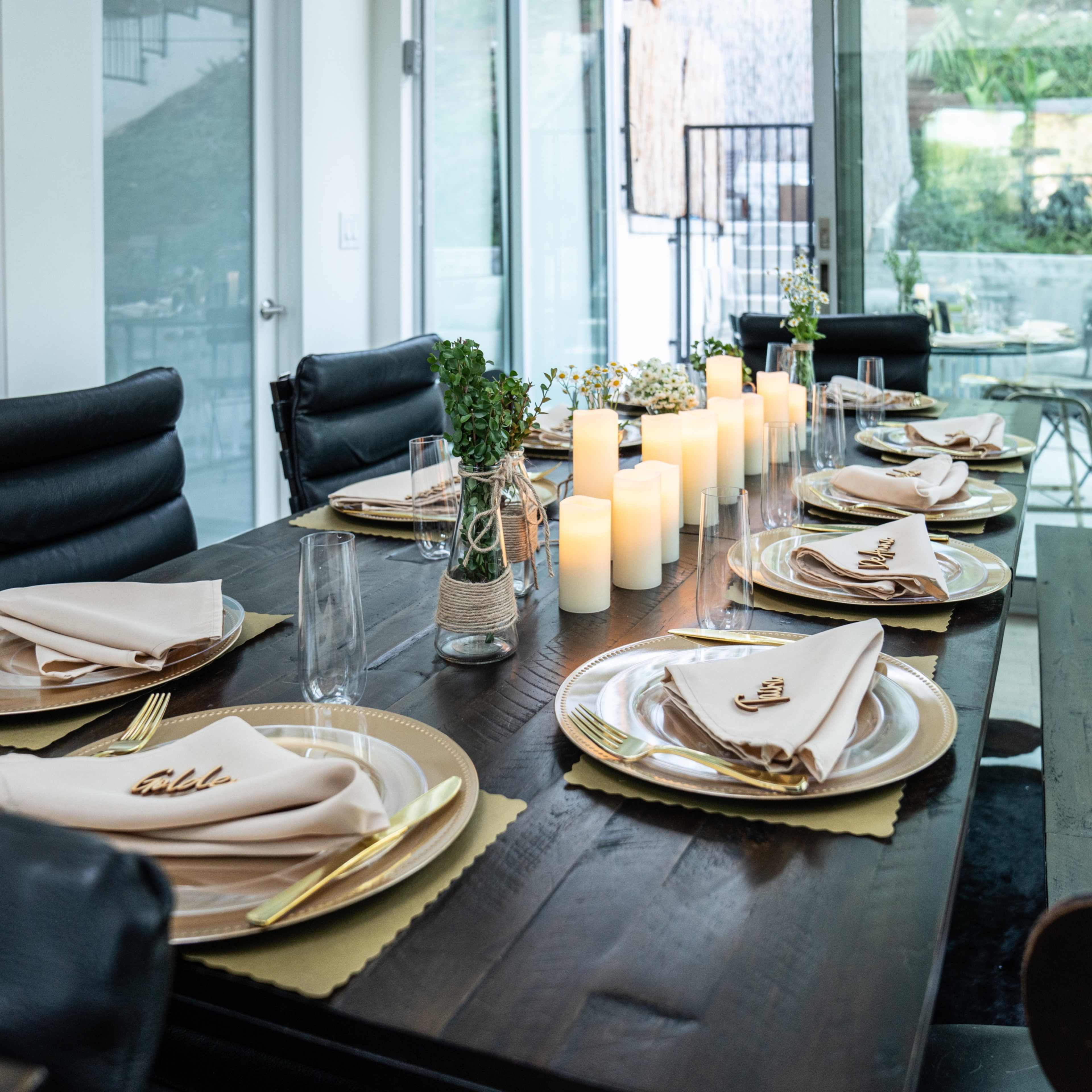 A long dining table is set with gold-bordered plates and neatly folded napkins, surrounded by chairs, with candles and potted greenery as centerpieces.