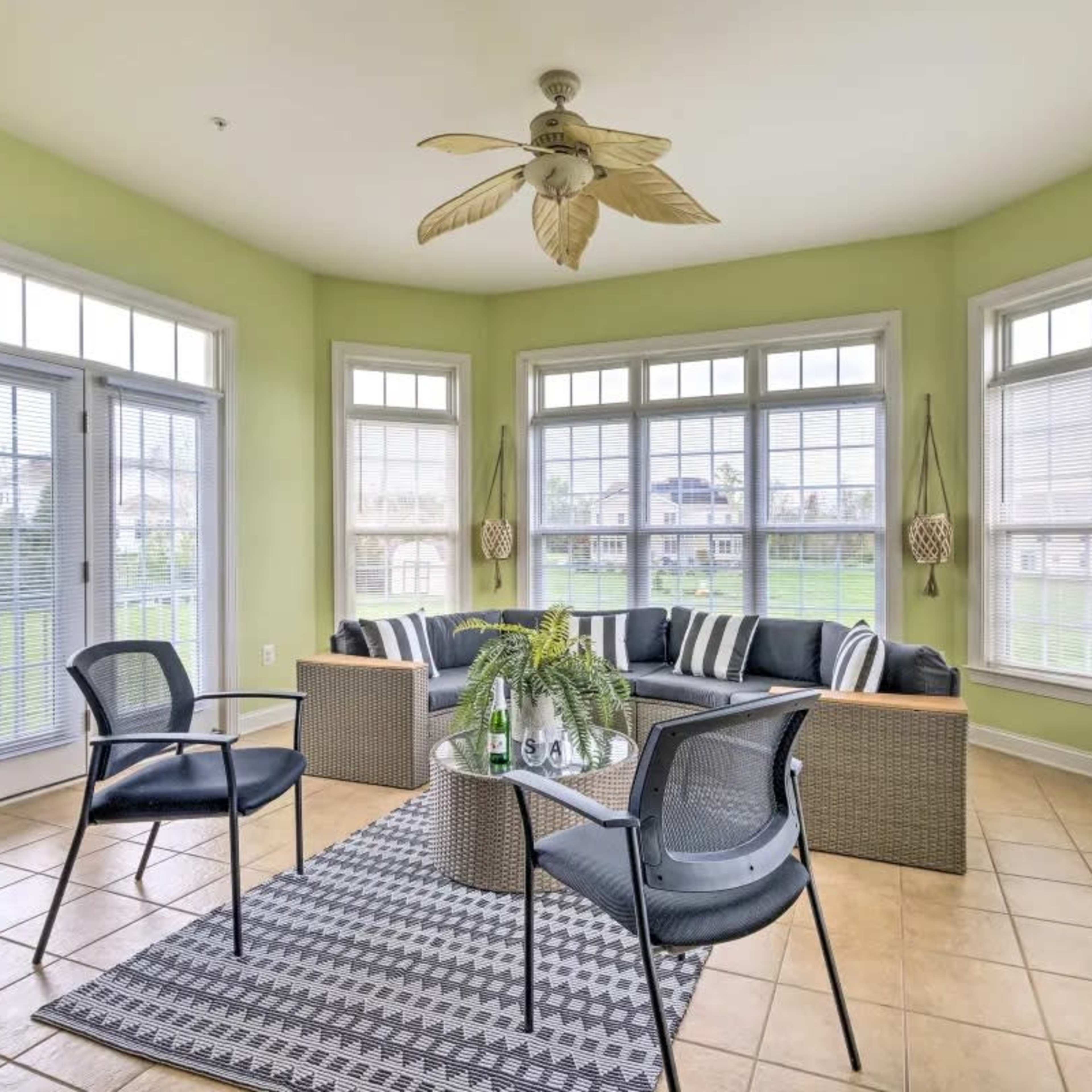 A sunlit sunroom features a rattan seating area with black cushions, surrounded by large windows and a patterned rug on tiled flooring.