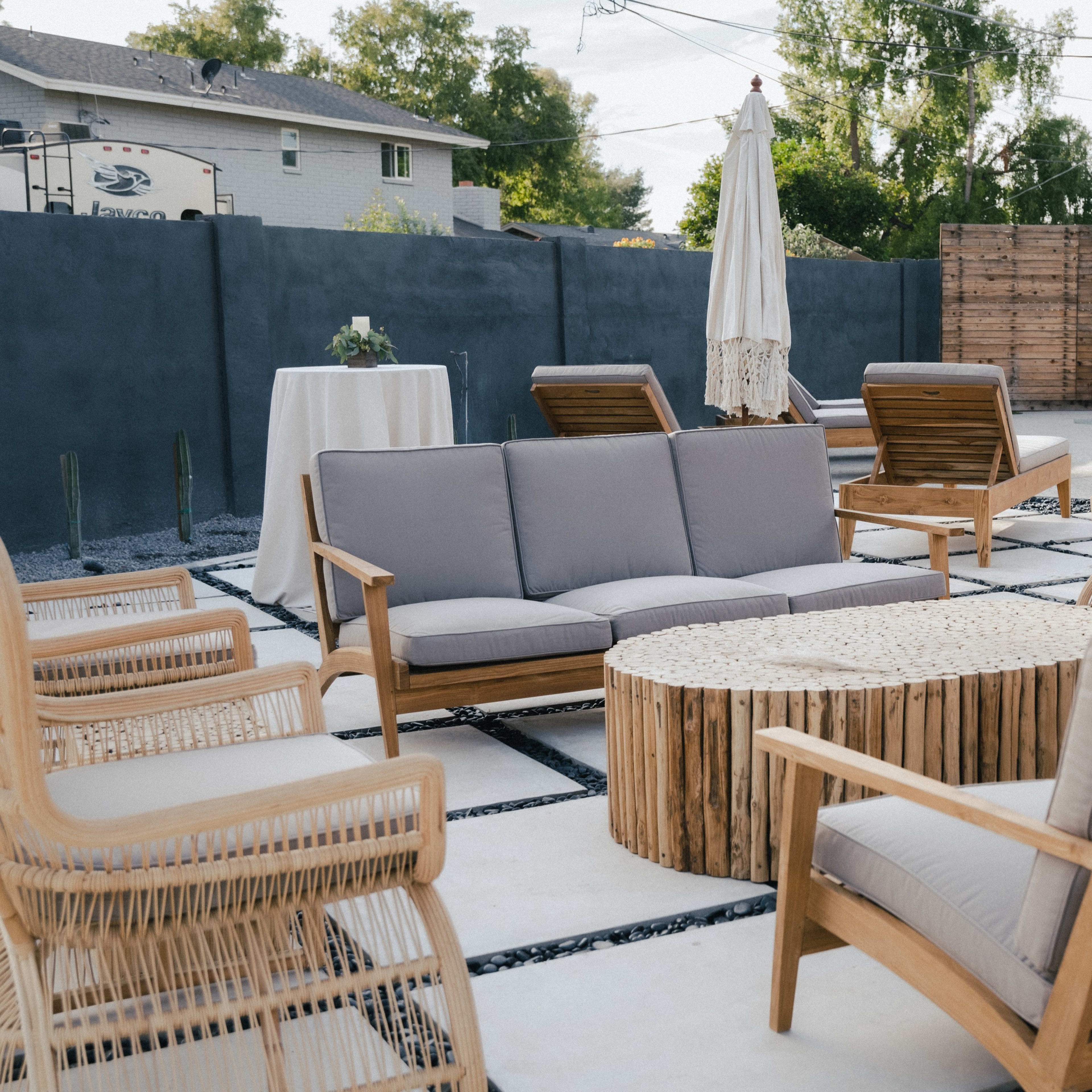 A patio area with wooden furniture, including a circular coffee table, surrounded by several chairs and two tall tables, in a setting with a pool and a wooden fence.