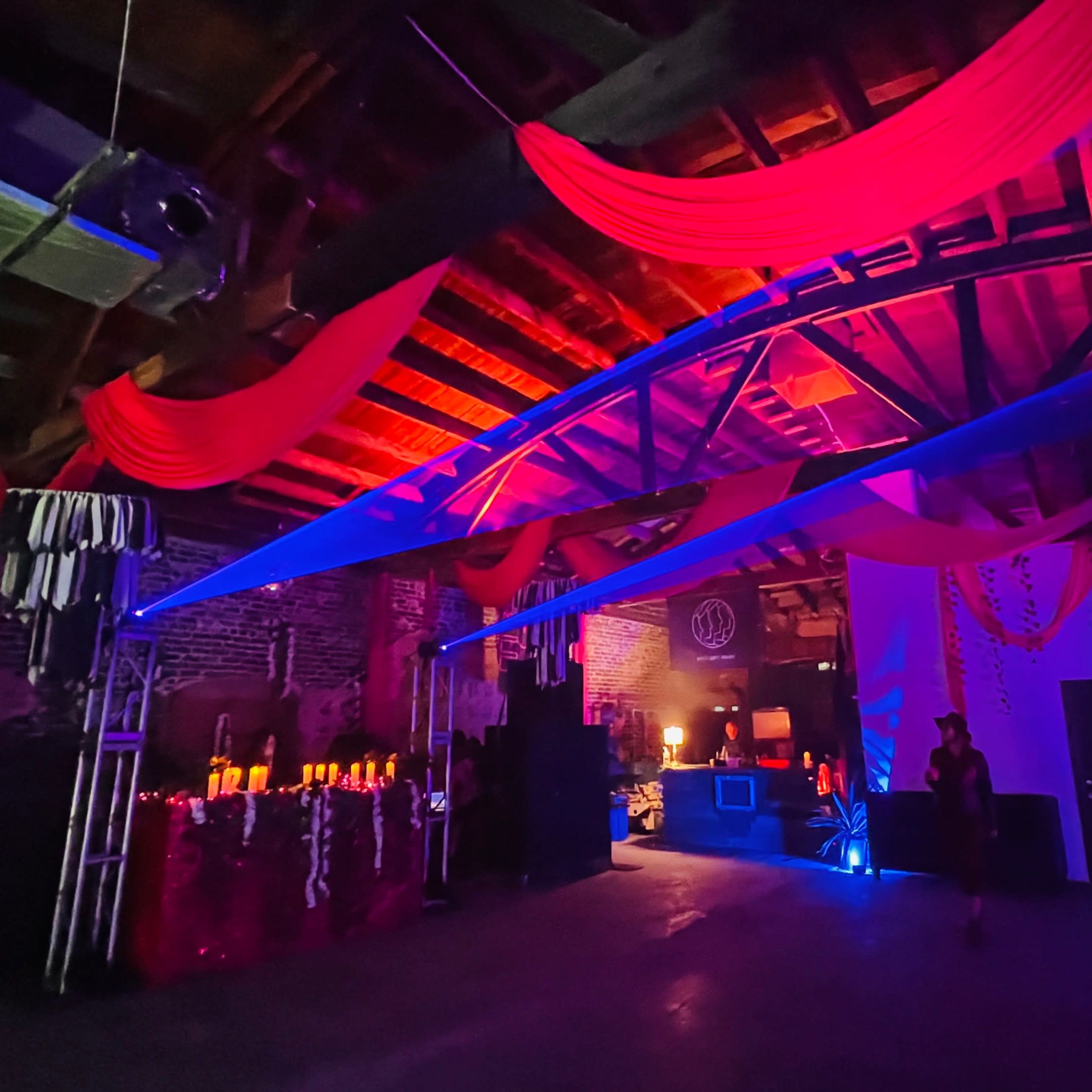 A dimly lit event space with red and blue drapery, soft lighting, and decorative elements, along with a DJ setup in the background.