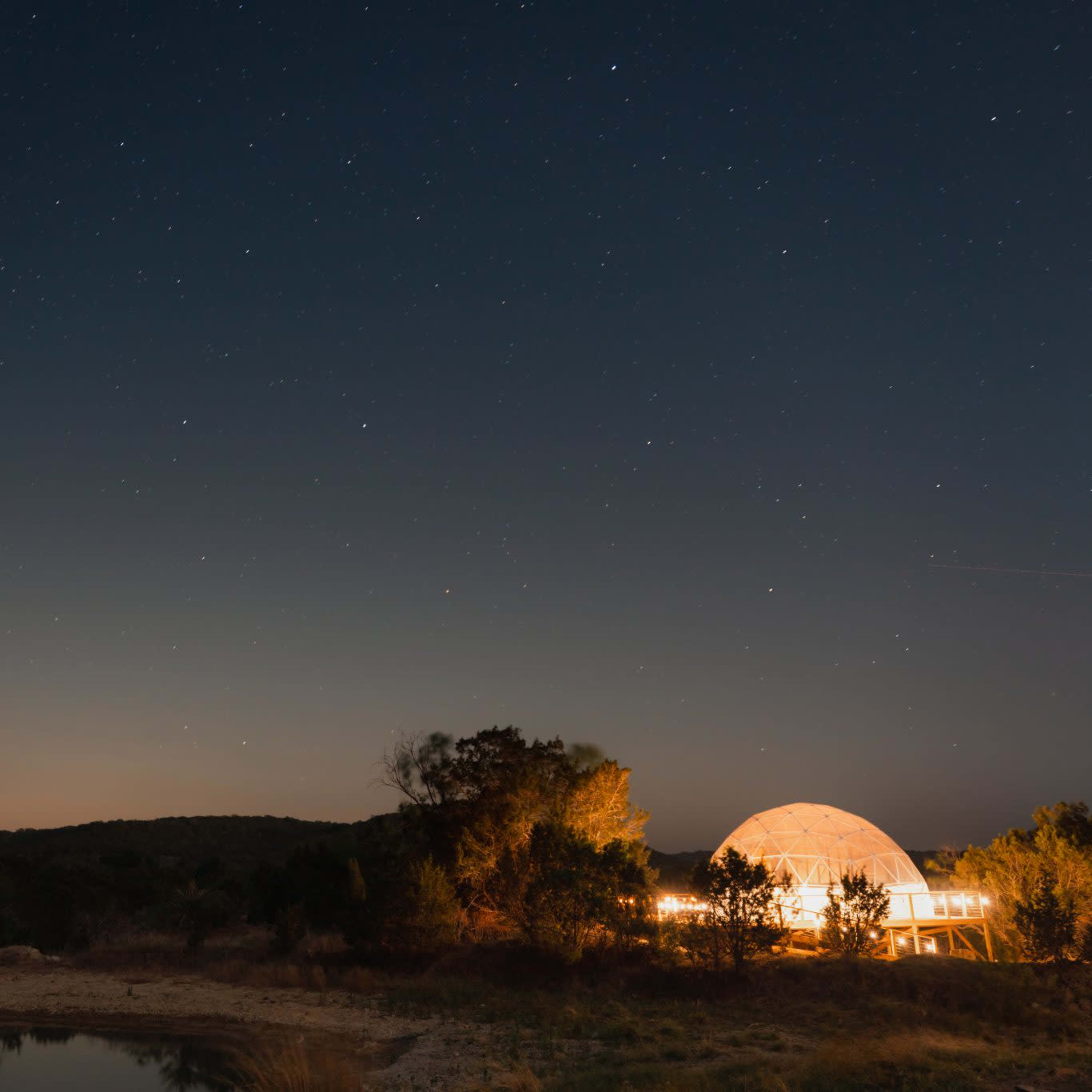 A dome-shaped structure is illuminated at night near a calm body of water, under a starry sky.
