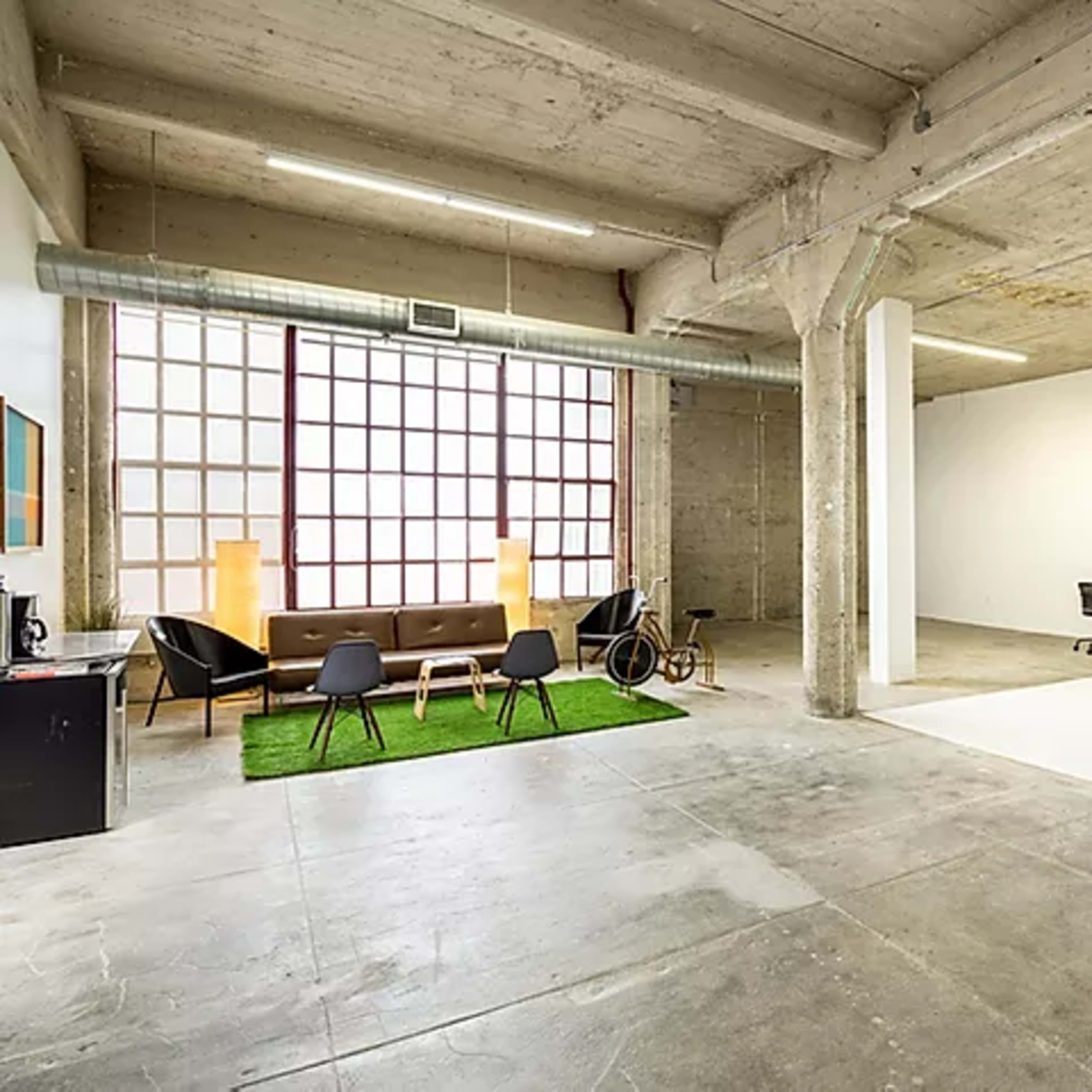 A spacious, industrial-style workspace featuring a seating area with a sofa and black chairs on green turf, adjacent to a bright, minimalist room with a desk and computer.