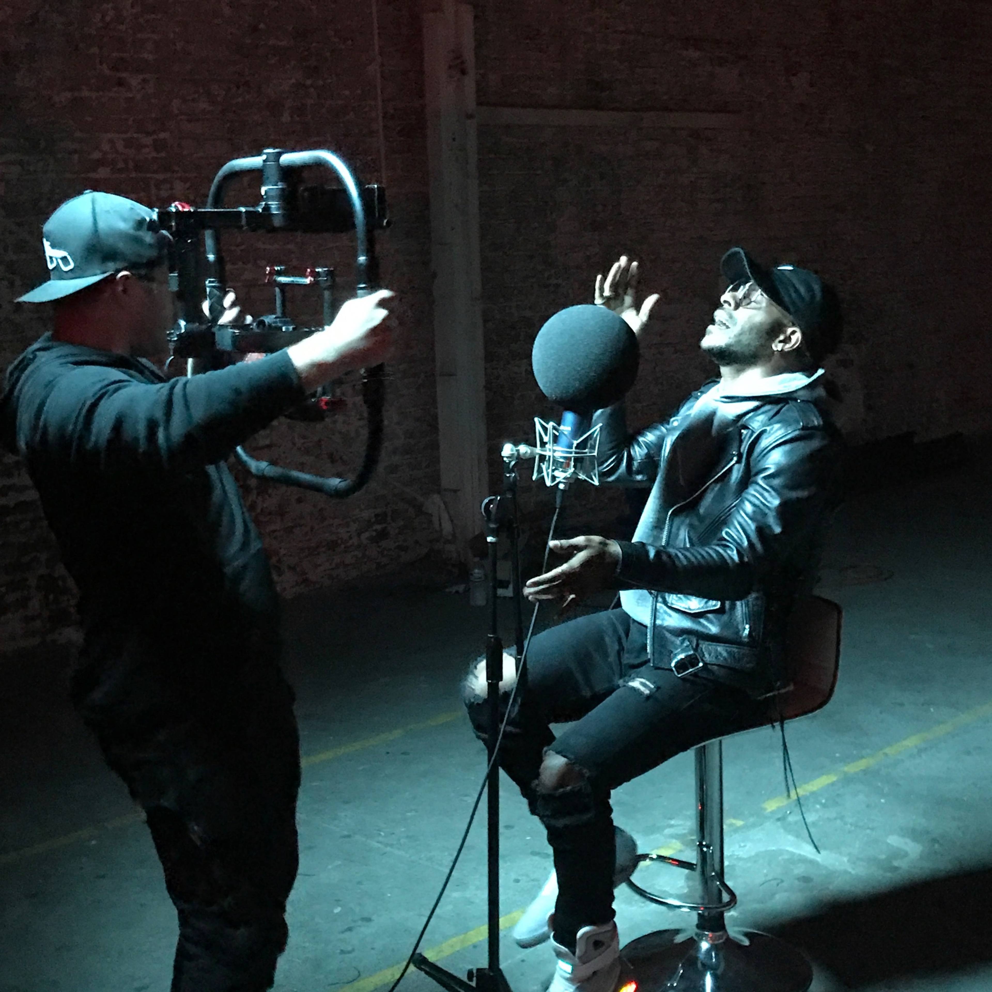 A man is seated on a stool in a dimly lit space while another person, holding a camera rig, films him as he interacts with a microphone.