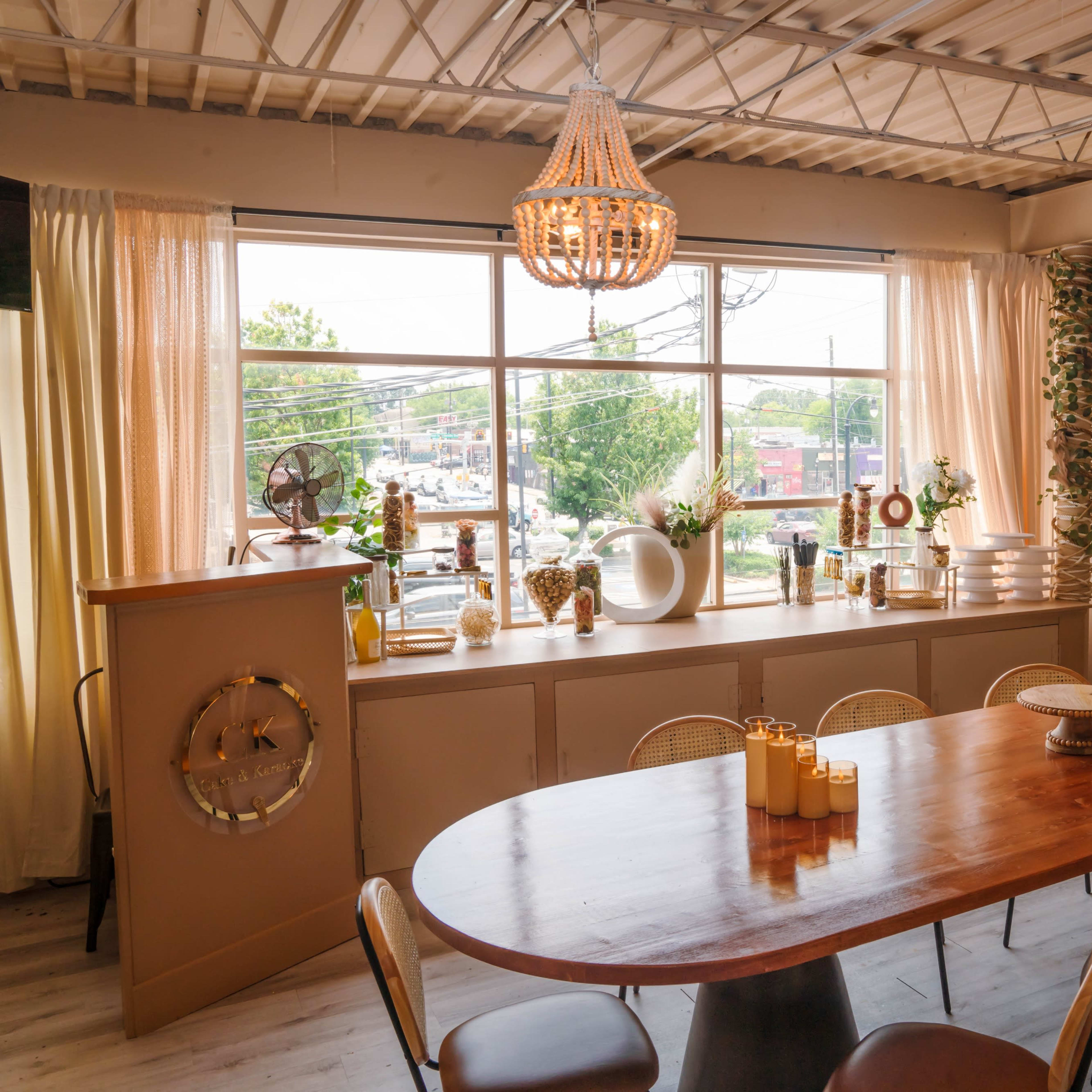 A spacious dining area features a long wooden table surrounded by chairs, with large windows allowing natural light to illuminate the decor and plants along the walls.