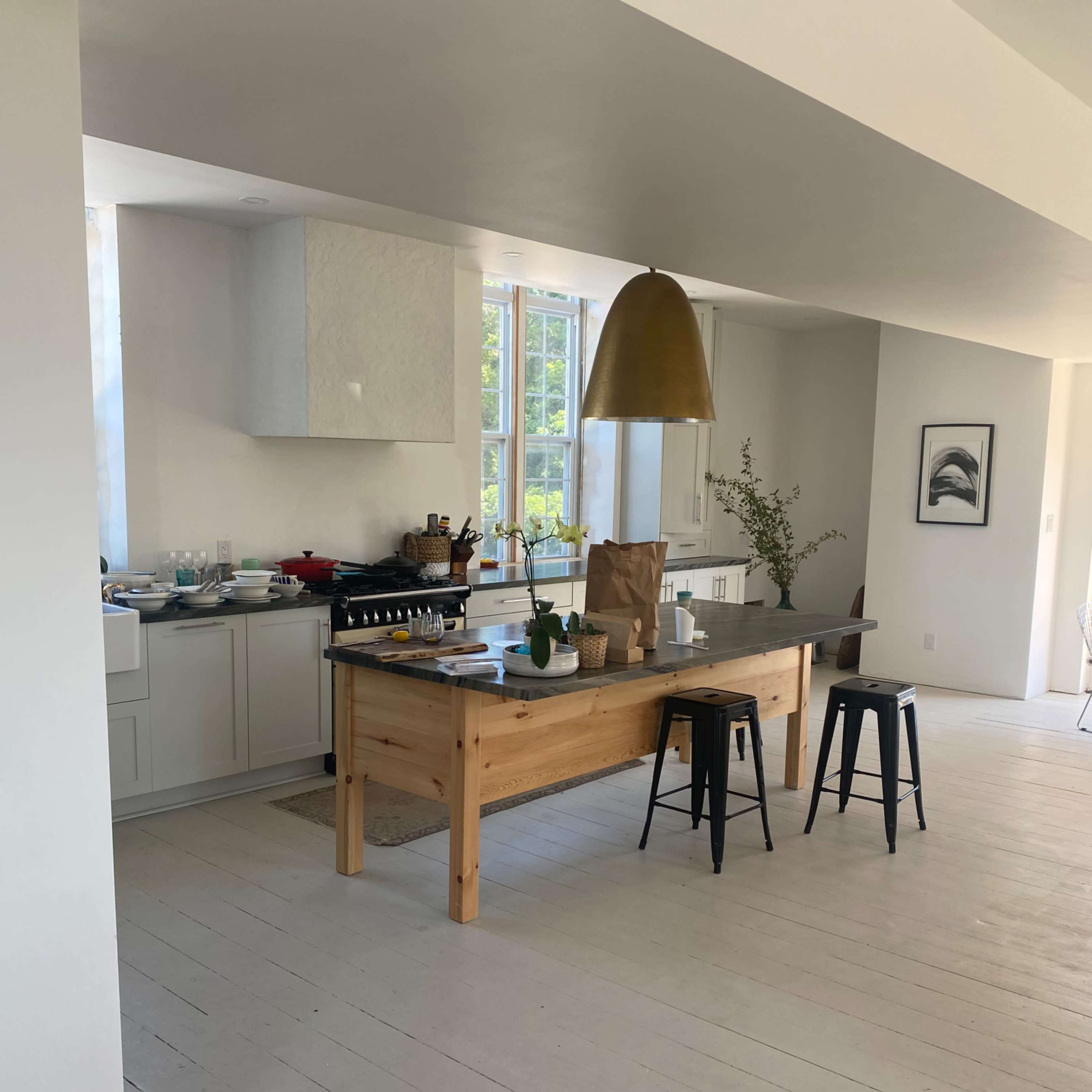 A modern kitchen features a wooden table with black stools, a gold pendant light overhead, and a clean, open design with white walls.