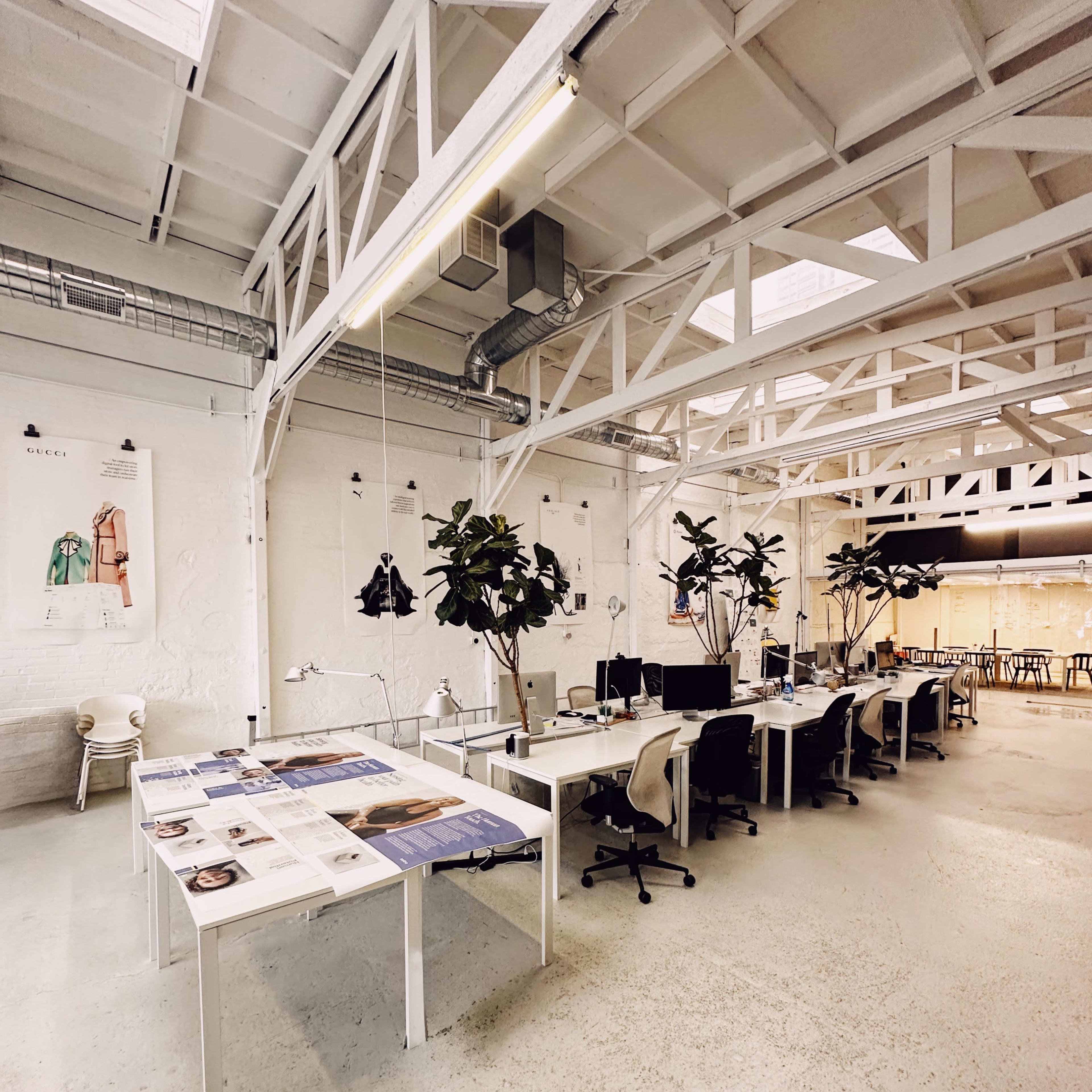 A spacious, modern office area with several workstations, large tables displaying printed materials, and greenery in the form of potted plants.