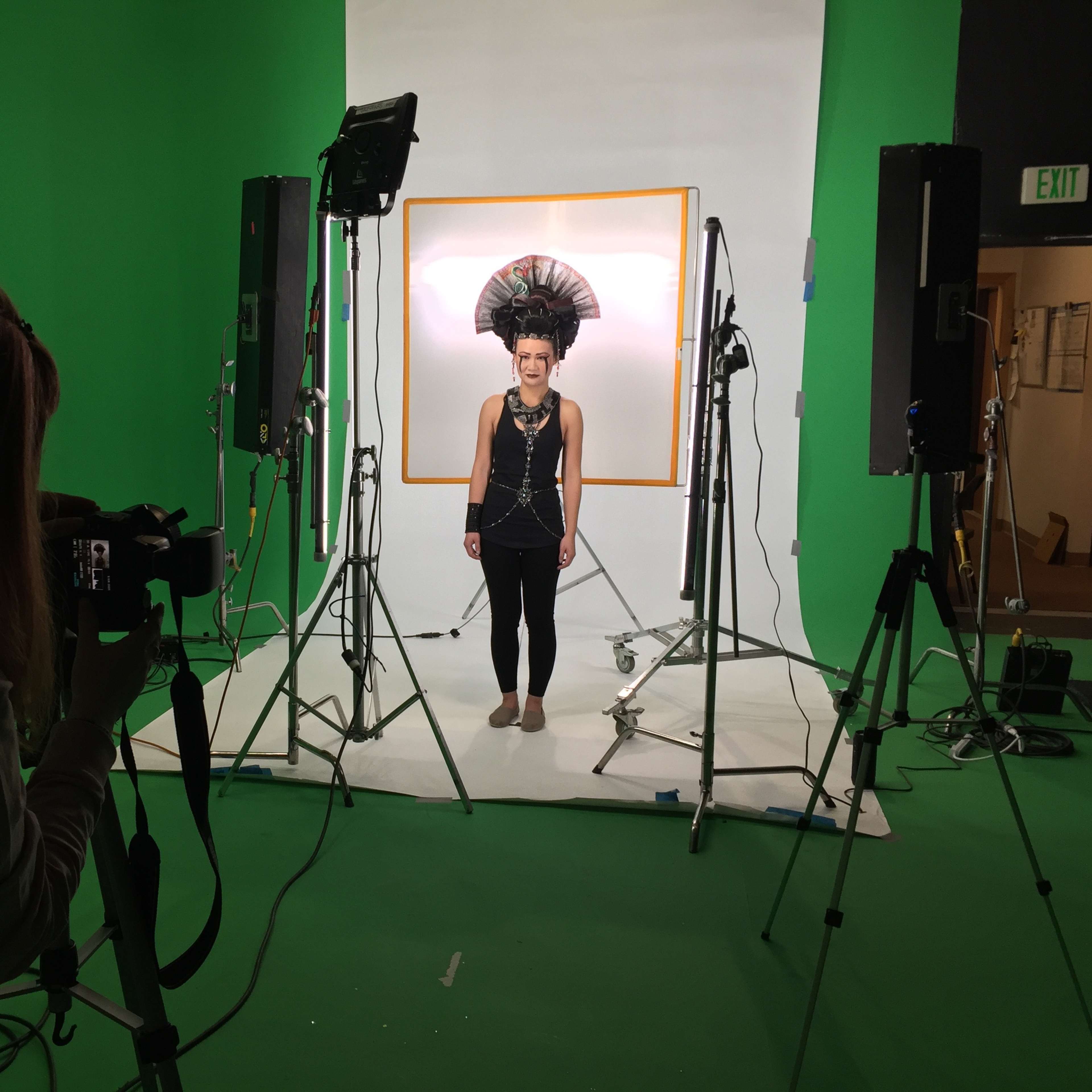 A person wearing a large feathered headdress poses for a photo in a studio with a green backdrop while another person takes the picture.