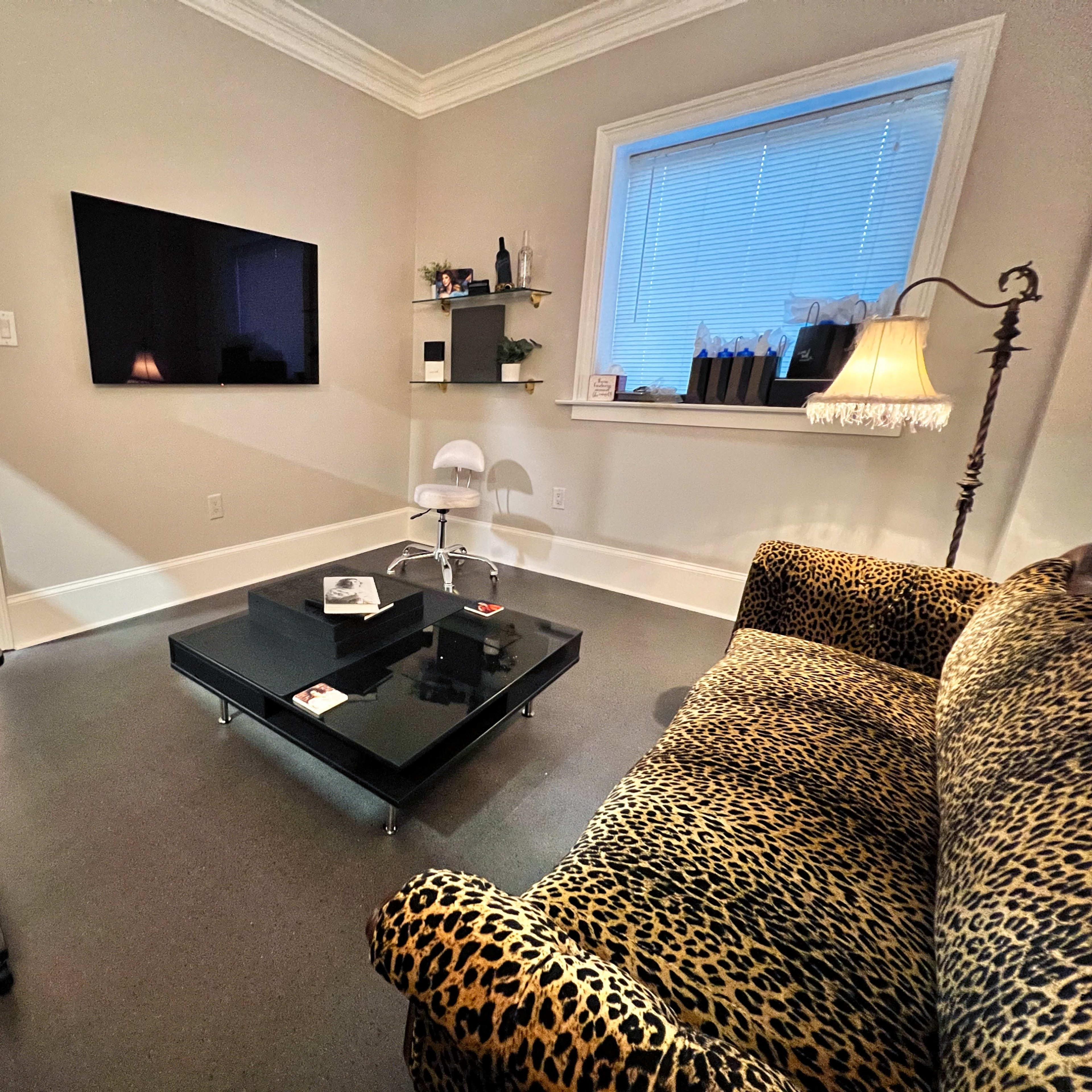 A modern living room with a leopard-print sofa, a black coffee table, a television mounted on the wall, and a lamp beside a window.